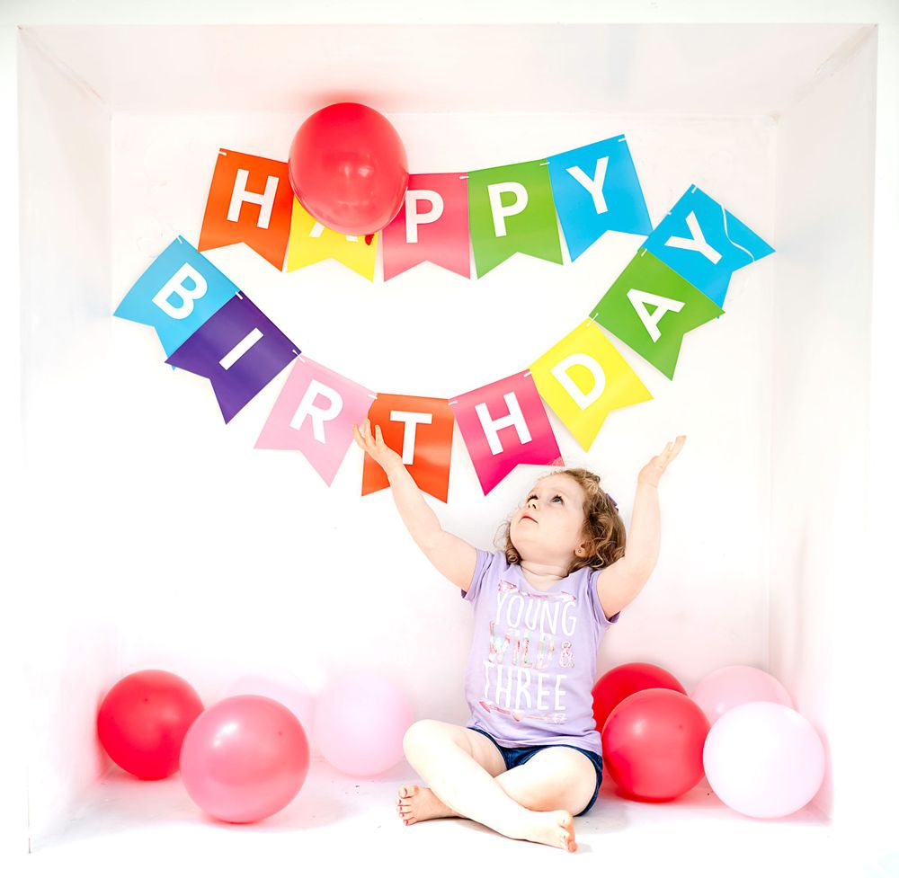 Birthday celebration setup with rainbow banner and red balloons against white background.