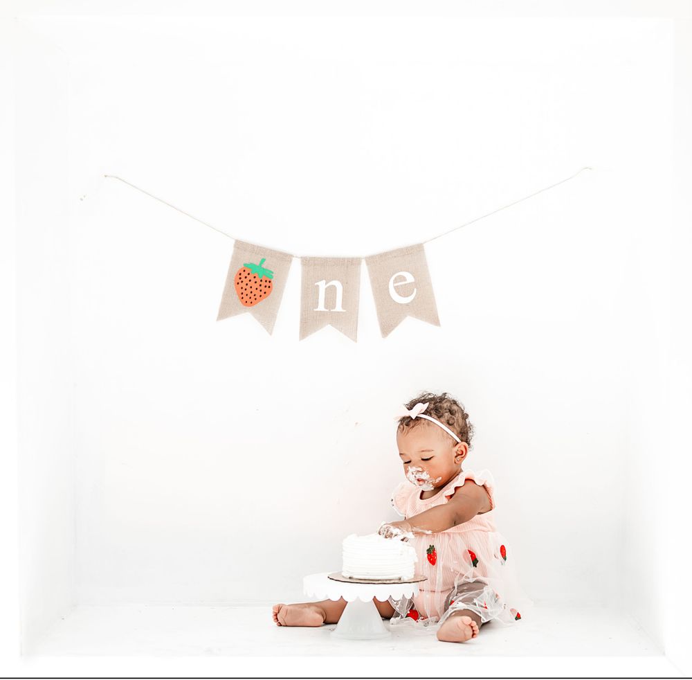 Simple first birthday cake smash setup with strawberry banner and white backdrop.