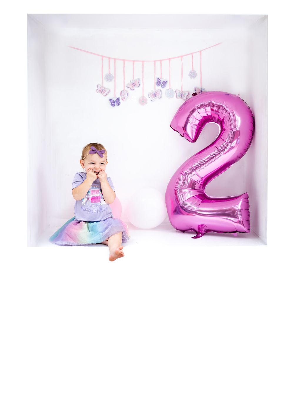 A second birthday photoshoot with a large pink number 2 balloon and butterfly decorations.