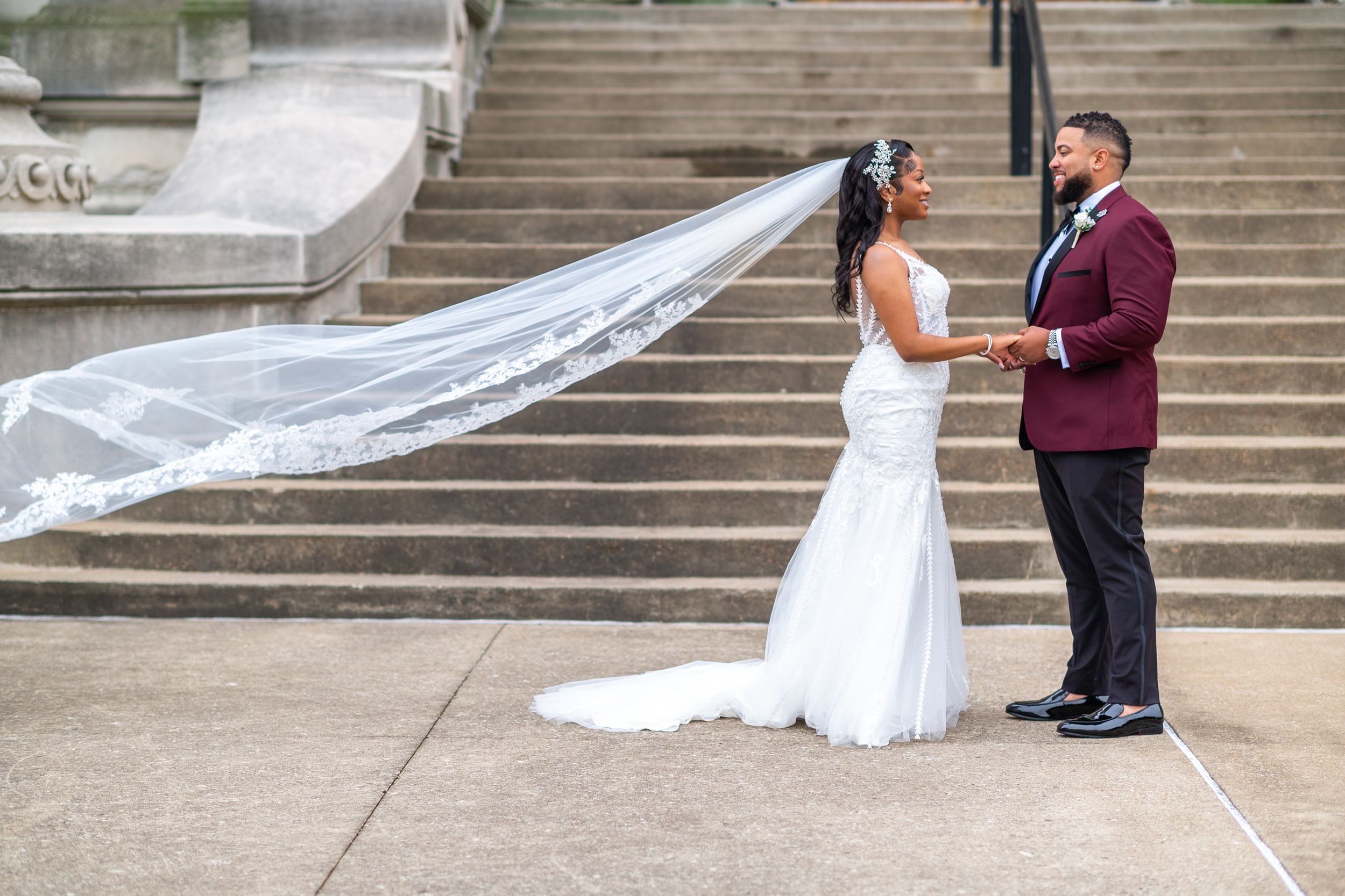 Husband and wife holding hands outside and looking into each other eyes, veil is stretched in the air and floating behind her head