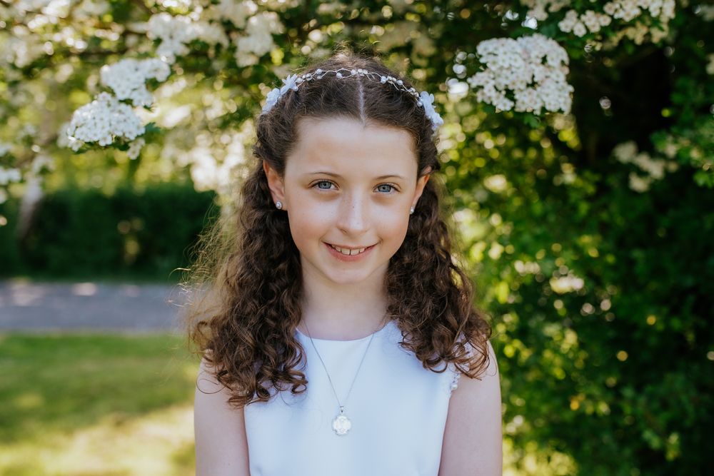 First Communion girl portrait outdoors, sunny morning light, soft green tree background, natural candid child photography, elegant communion
