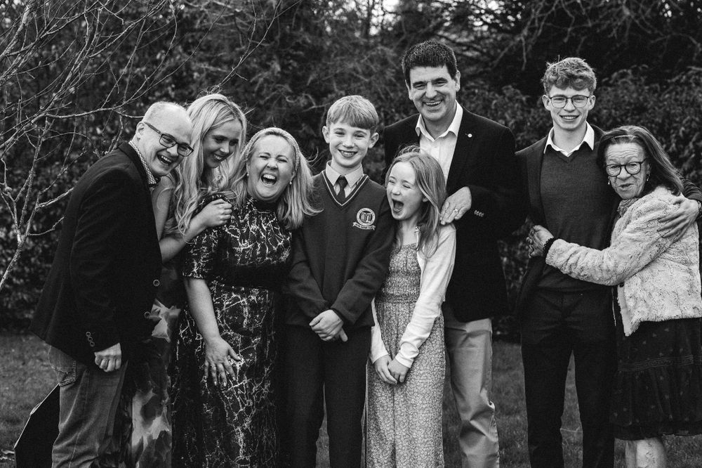 A group of seven people pose together outdoors in a black and white image, surrounded by trees. They display a range of cheerful expressions