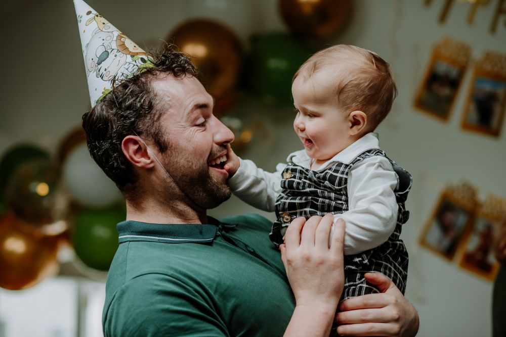 A person in a party hat holds a young child dressed in a plaid outfit, surrounded by festive decorations and balloons.