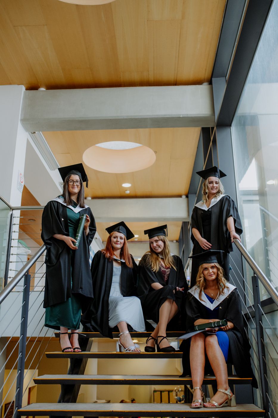 A group of graduates in caps and gowns poses on a staircase, holding diplomas and smiling, celebrating their achievement.
