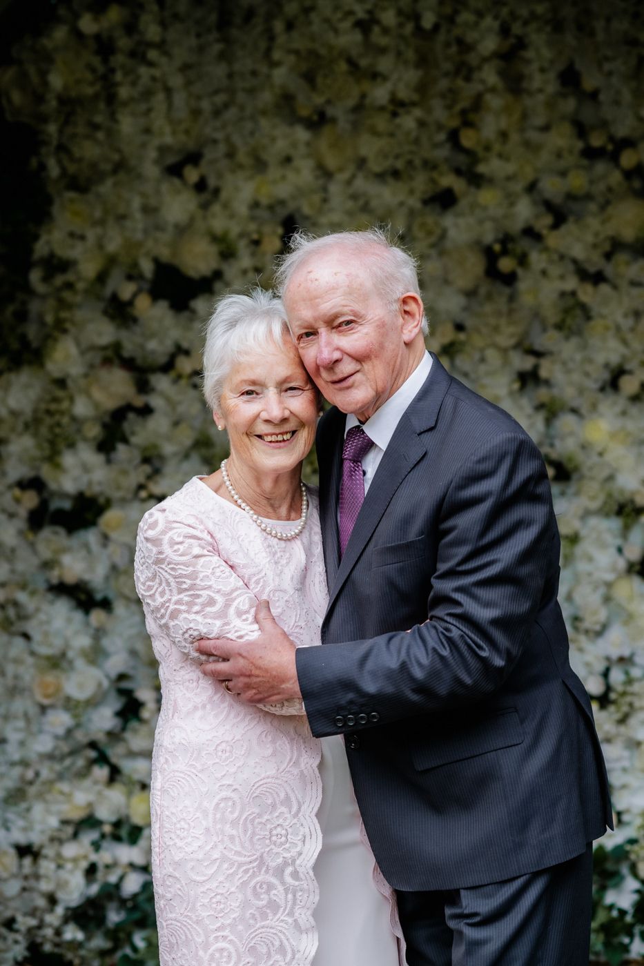 A couple embraces, dressed elegantly against a beautiful floral backdrop, radiating joy and love at a special event.