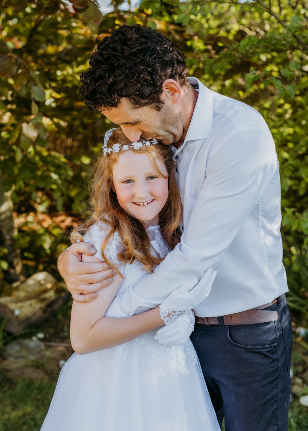 A joyful moment shared between a child in a white dress and an adult, surrounded by lush greenery in a serene outdoor setting.