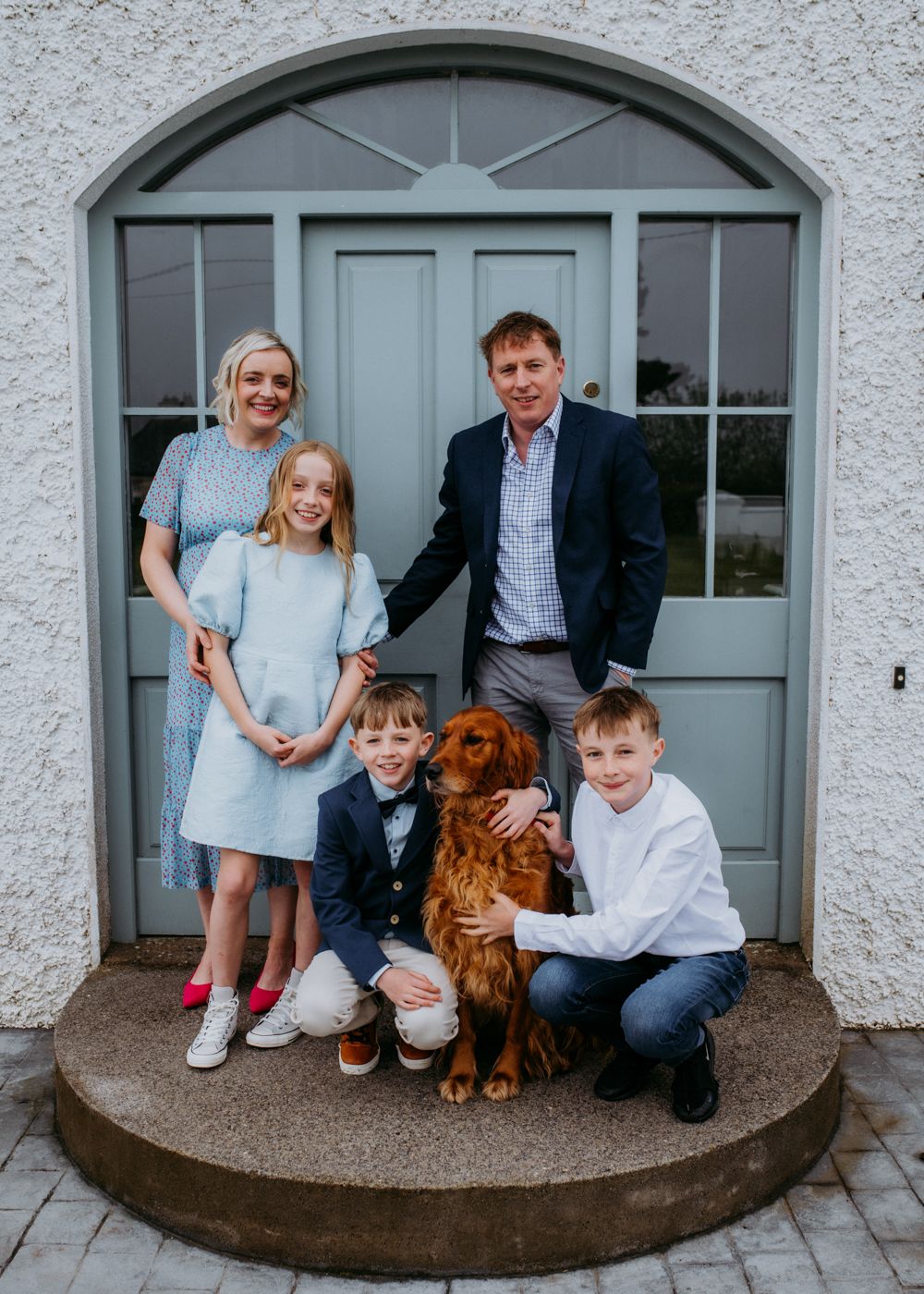 A family poses on a porch with a golden retriever, dressed in smart casual outfits, showcasing a warm and cheerful atmosphere.