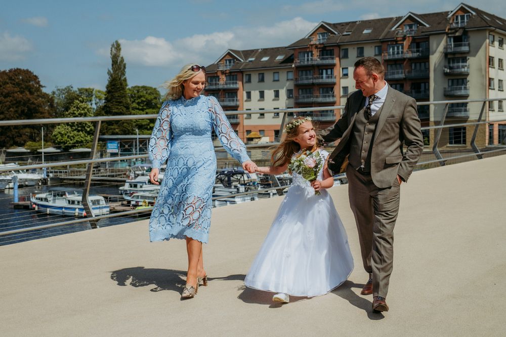 A family walks hand in hand across a new bridge in Athlone on a sunny day. The young girl, wearing a white dress and holding a small bouquet