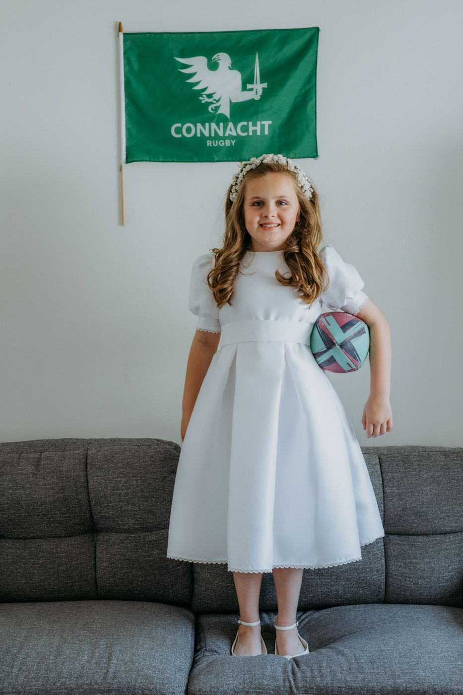 A girl in a white dress stands on a couch, holding a colorful bag, with a Connacht Rugby flag hanging on the wall behind her.
