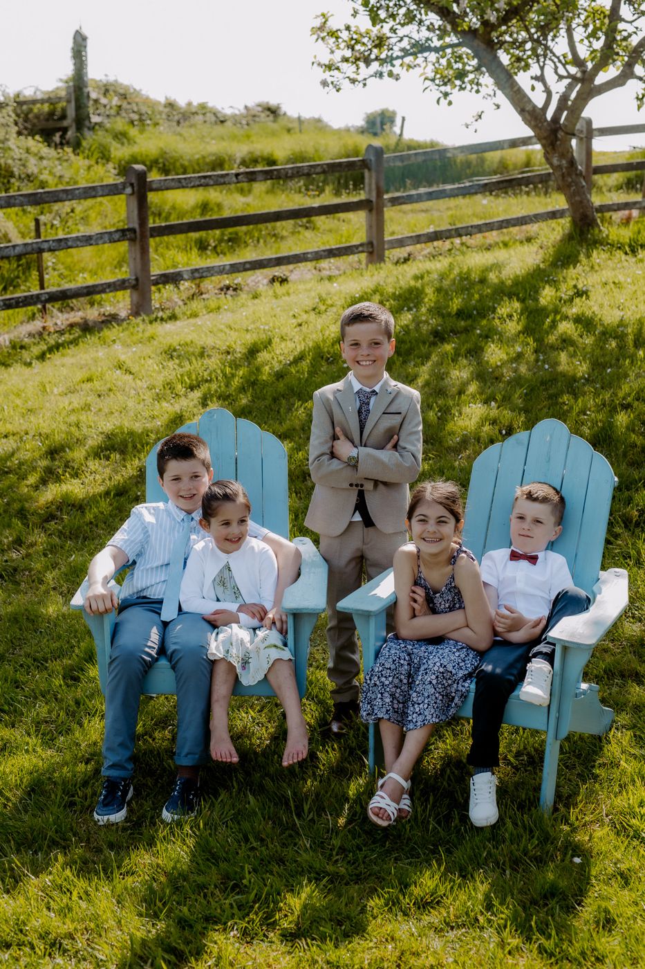A group of five children dressed smartly, sitting in blue chairs on a sunny day, surrounded by green grass and trees.