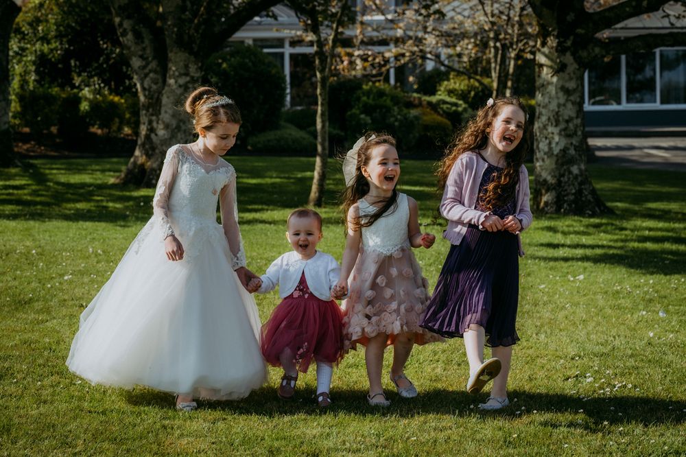 Four girls in colorful dresses walk together on a green lawn under sunny skies, surrounded by trees and a garden backdrop.