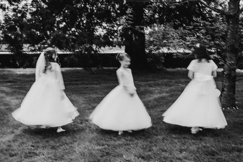 Three girls in white dresses twirl gracefully in a grassy area, surrounded by trees, captured in a black and white photograph.