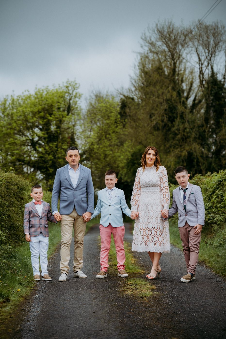 A family stands together on a country lane, dressed in stylish outfits, surrounded by lush greenery and overcast skies.