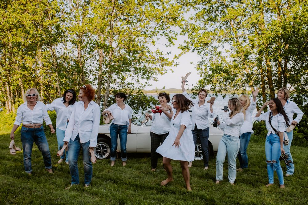 A lively group of women in white tops joyfully celebrating outdoors near a vintage car, surrounded by greenery and water.
