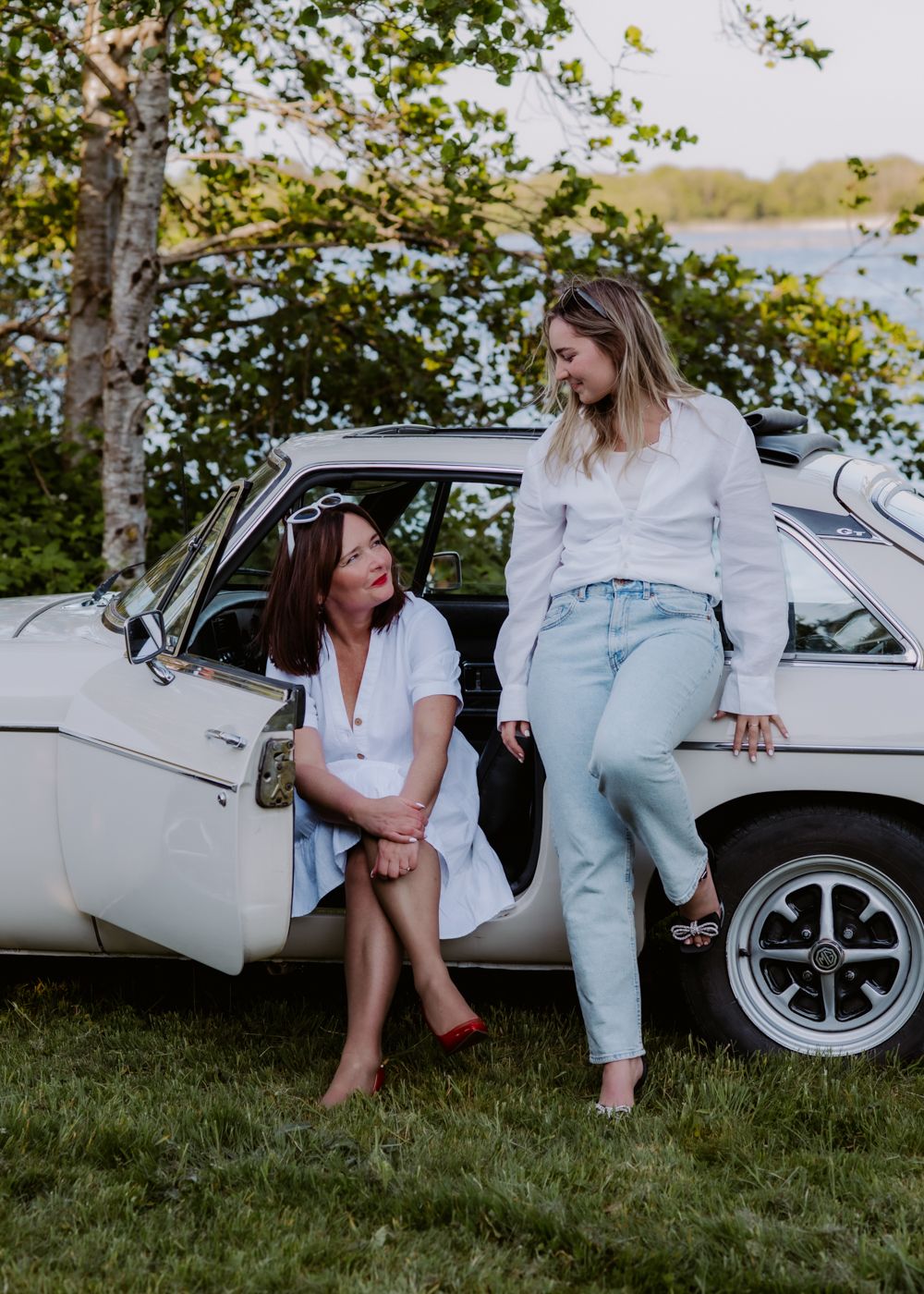 Two women lounge by a vintage white car near a lush lakeside, surrounded by greenery and soft sunlight.