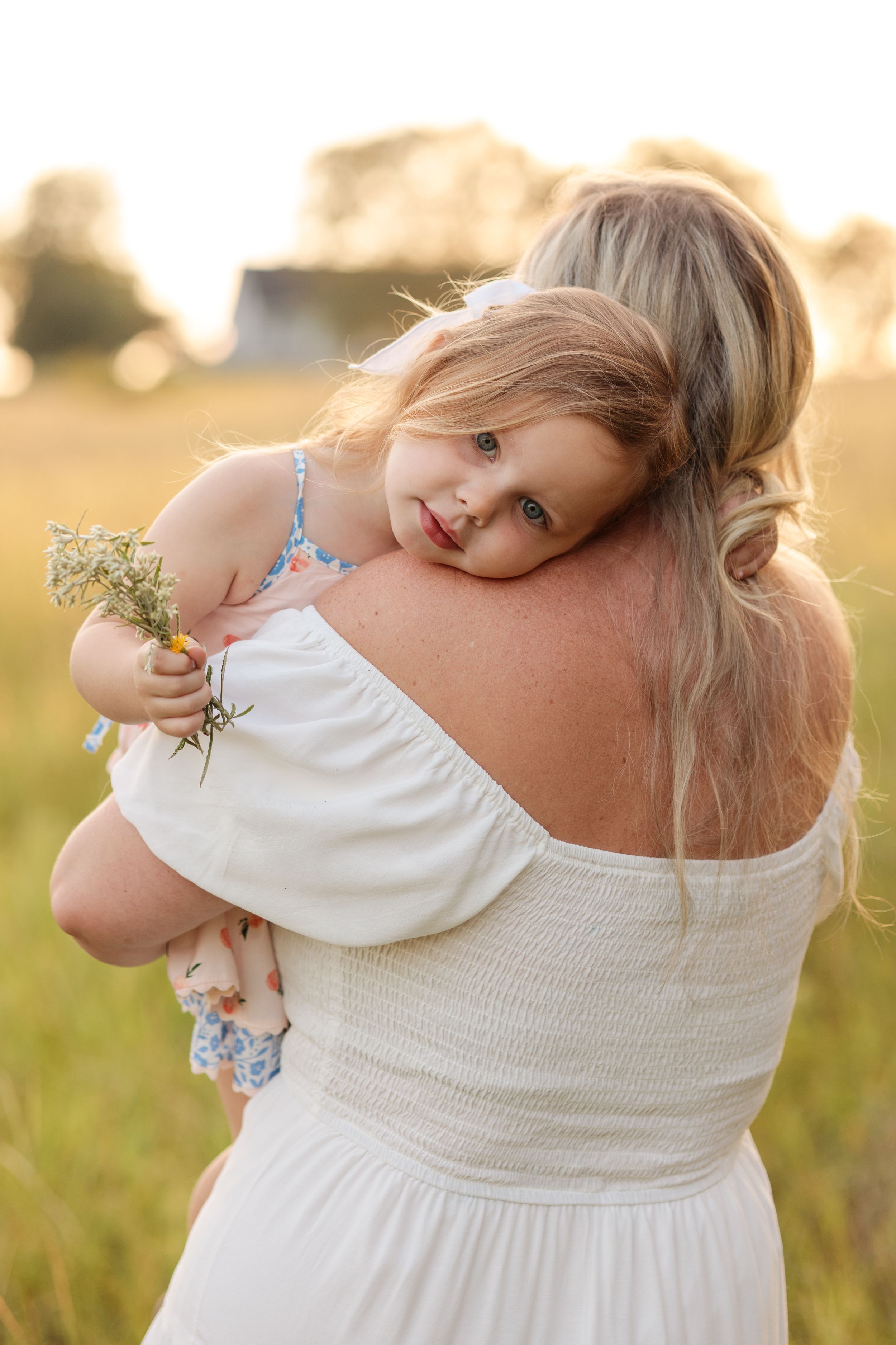 A tender moment captured at golden hour as a child in white holds wildflowers while being carried in a grassy field.