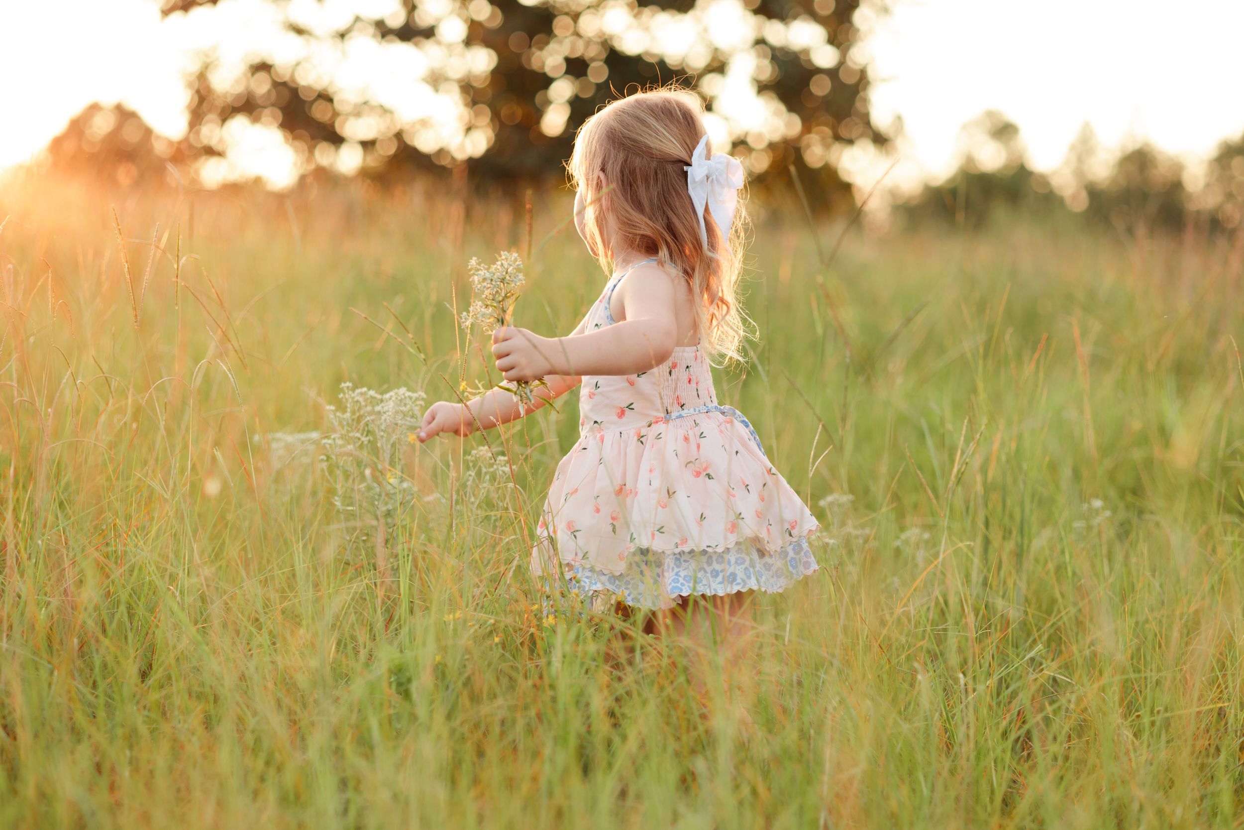 A figure in a white floral dress twirls gracefully in a golden sunlit meadow during sunset.