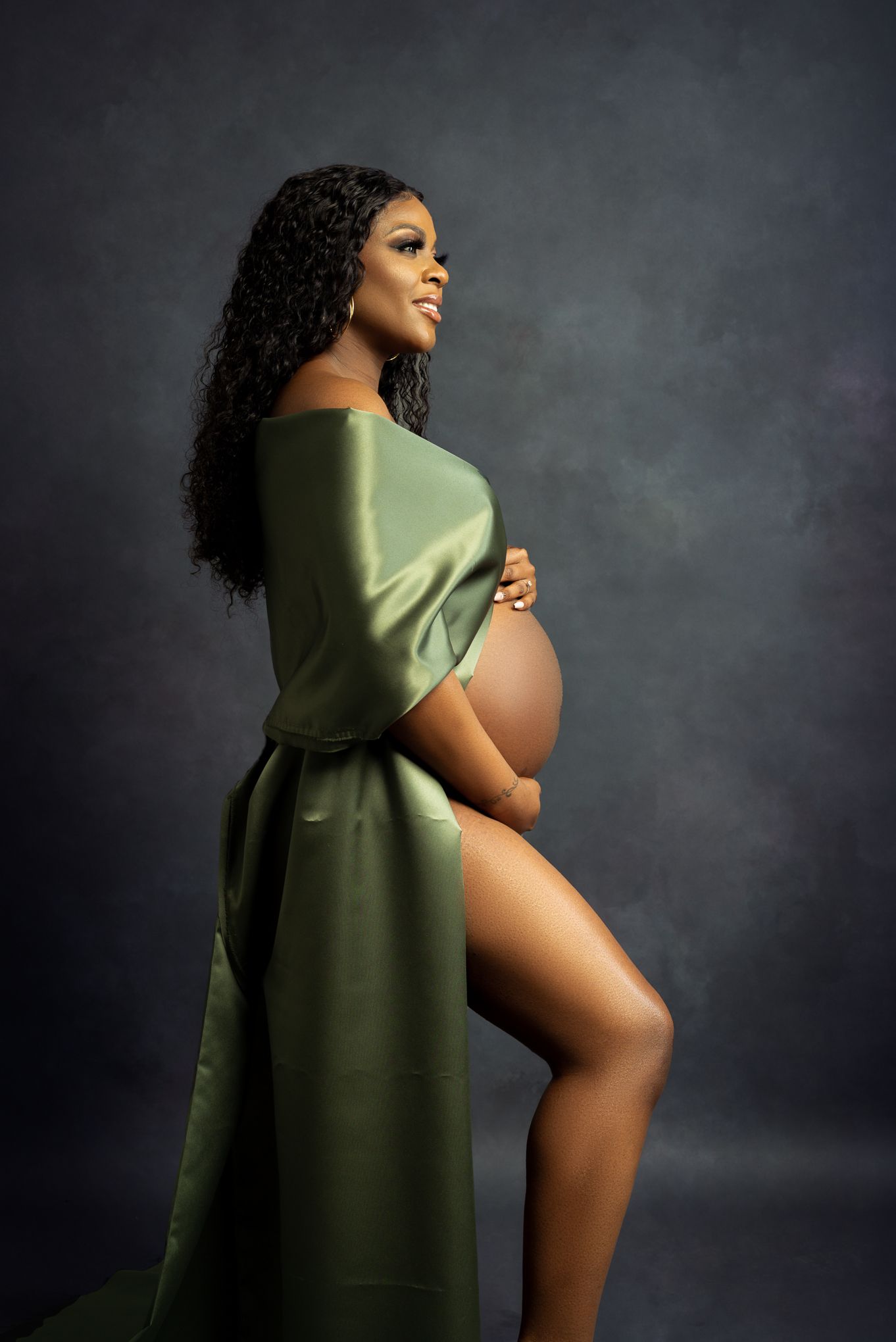 Artistic maternity portrait with green silk wrap against dark moody backdrop shows elegant silhouette in profile.
