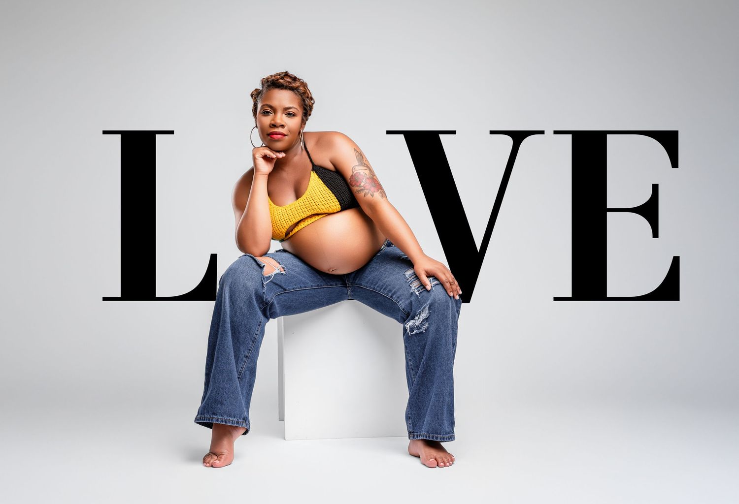 Pregnant woman in yellow top and jeans sits on stool against LOVE typography backdrop in maternity photo.