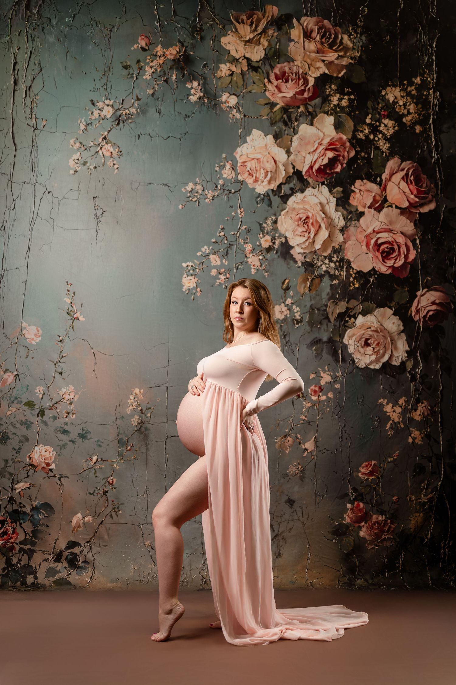 A maternity photo featuring a flowing pink dress against a vintage floral backdrop with pink roses and textured wall.