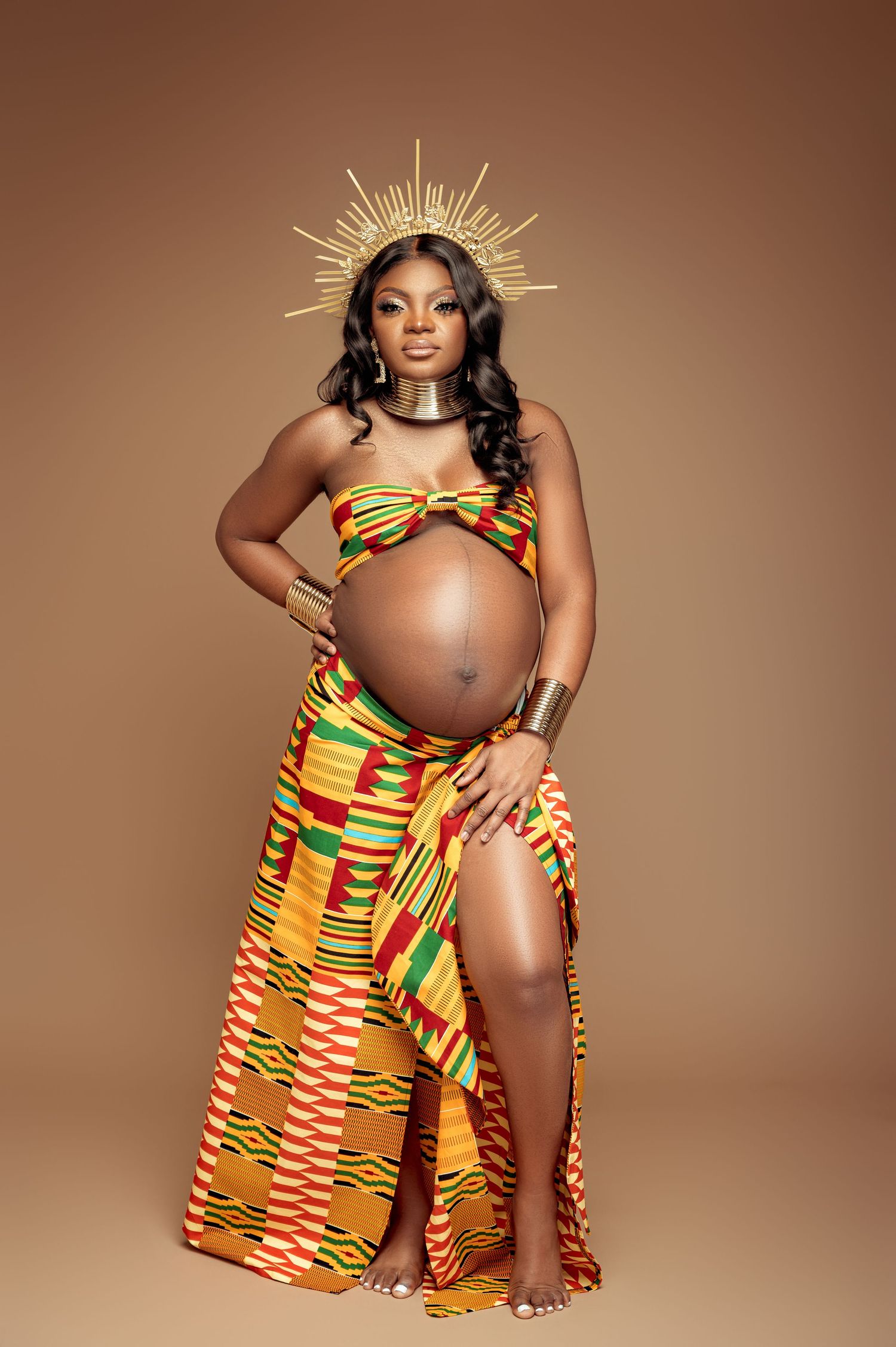 Maternity photoshoot featuring traditional African kente print dress in vibrant yellow, red and green patterns against brown backdrop.