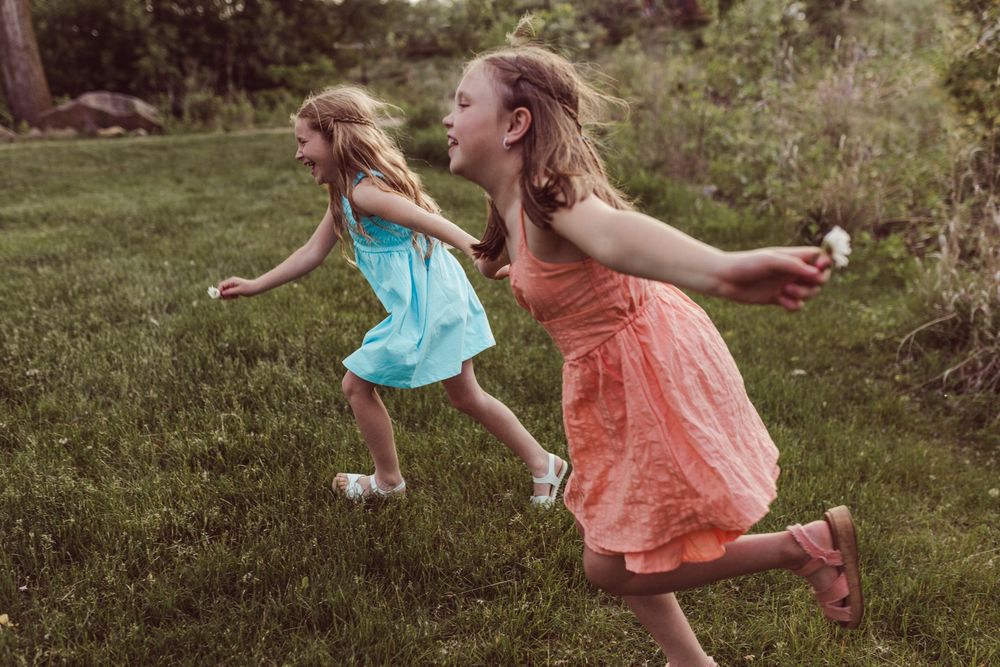 Two people in flowing dresses run playfully through a green garden on a summer day.