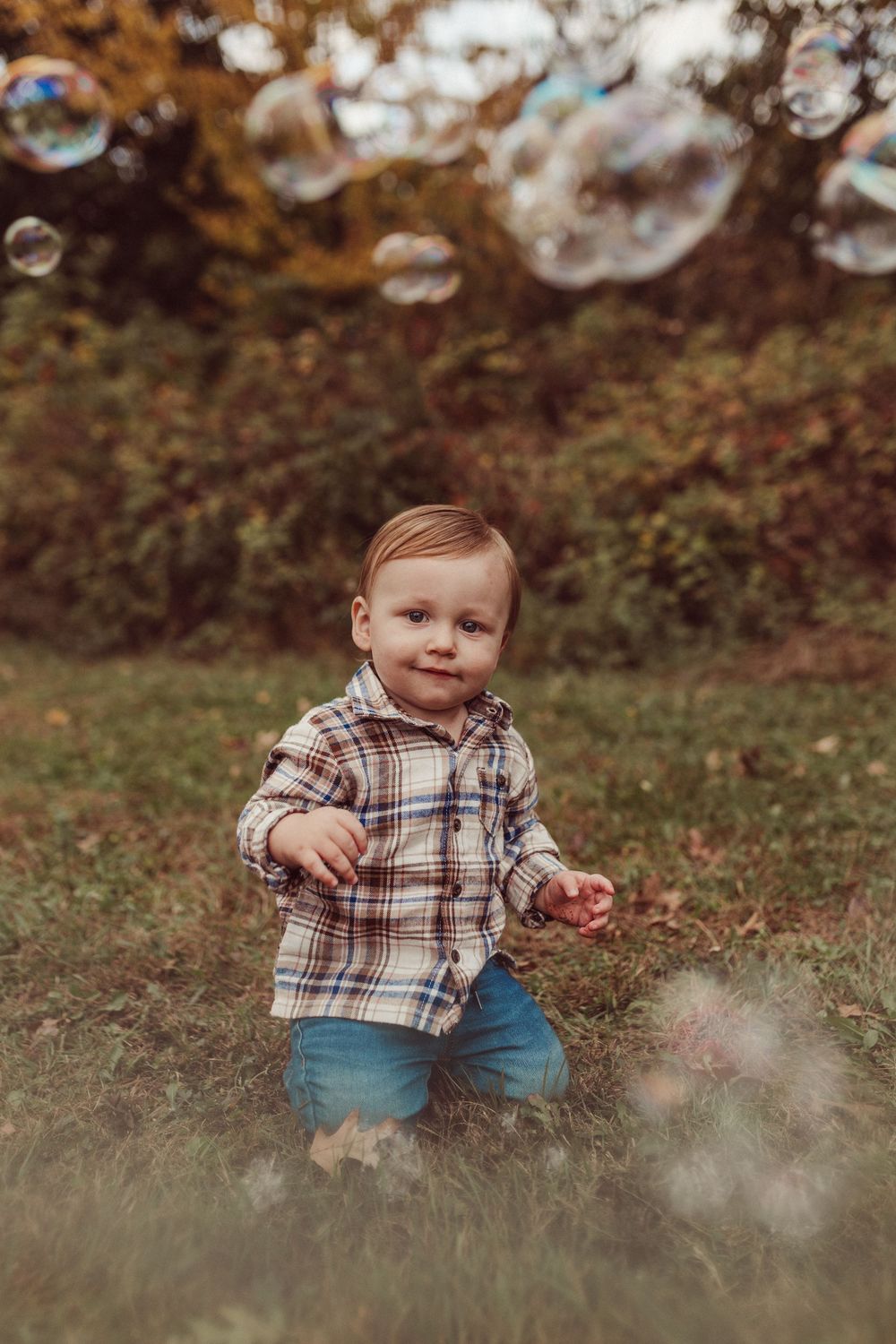 A small child in a plaid shirt and jeans sits in a grassy field during autumn.