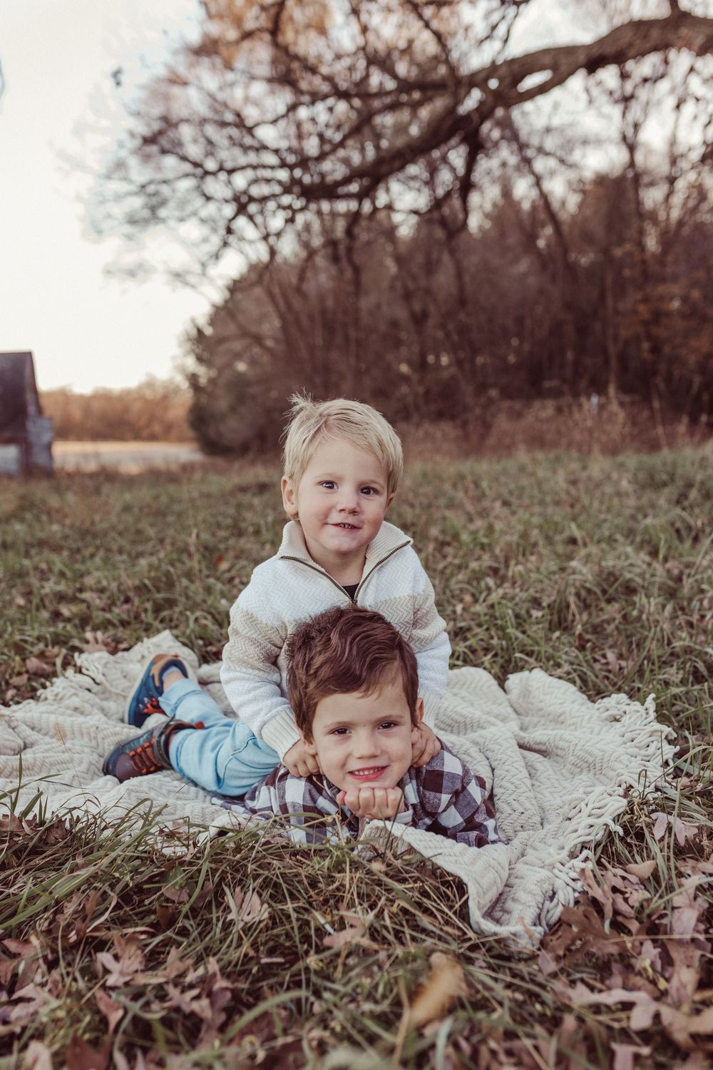 Two children pose playfully on a white blanket outdoors during autumn with fallen leaves and trees in the background.
