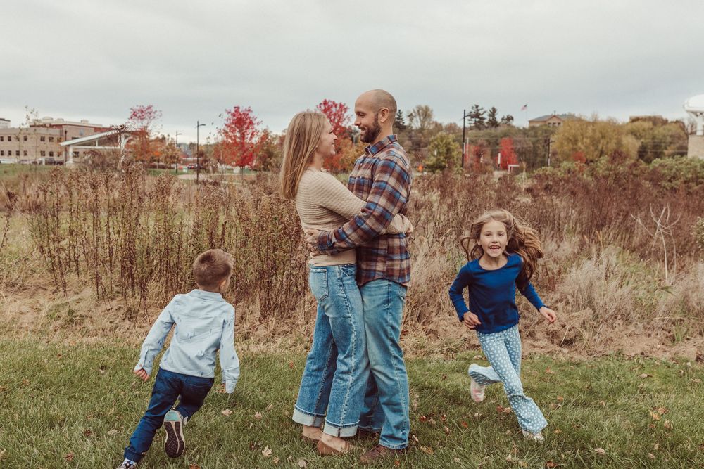 A family enjoys a playful moment together in an autumn field with tall grass and trees in the background.