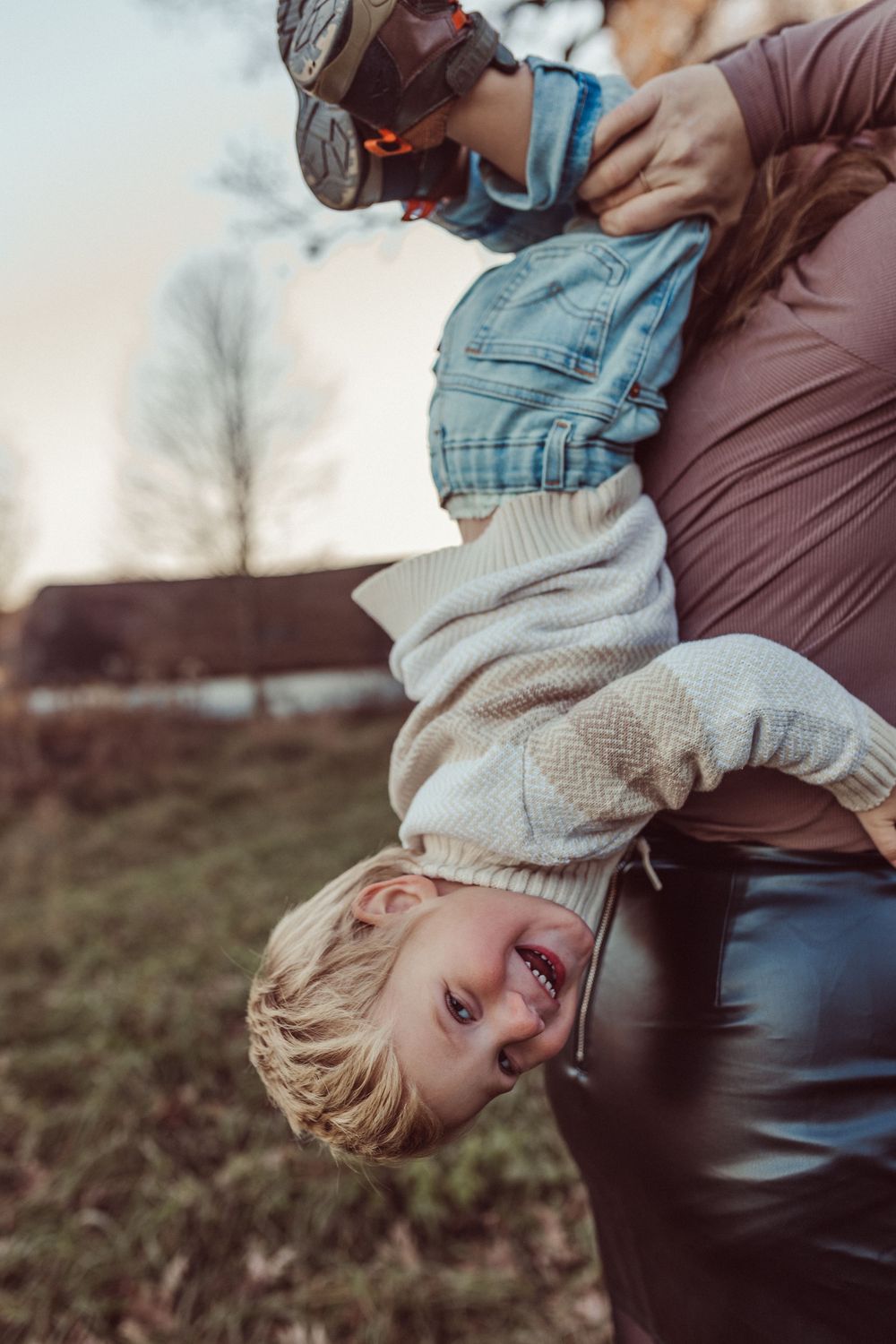 A child in overalls and sweater is held upside down while playing and laughing outdoors.