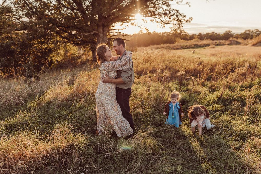 A couple embraces in a golden field at sunset with a toddler and dog beside them.