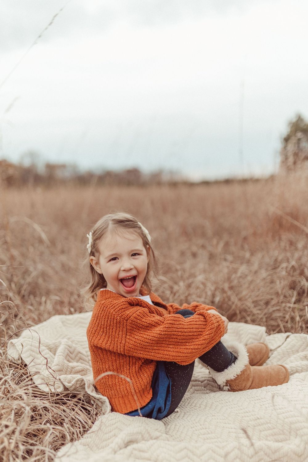 Child wearing orange sweater playing and rolling on cream blanket in autumn field.