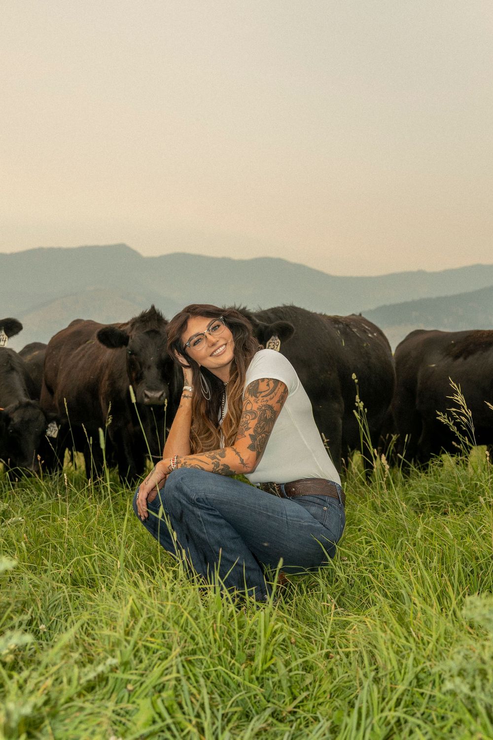 A smiling tattooed woman in jeans crouches among black cattle on a green hillside with mountains in the background.