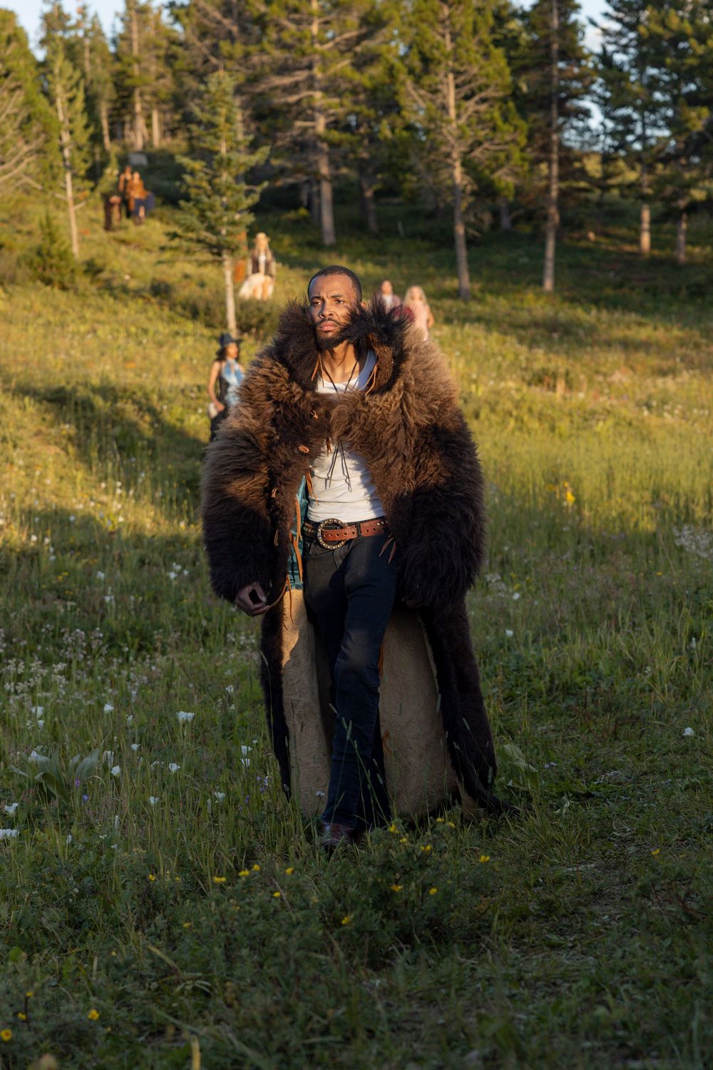 Someone wearing a black faux fur coat plays in a sunny mountain meadow with wildflowers and evergreen trees.
