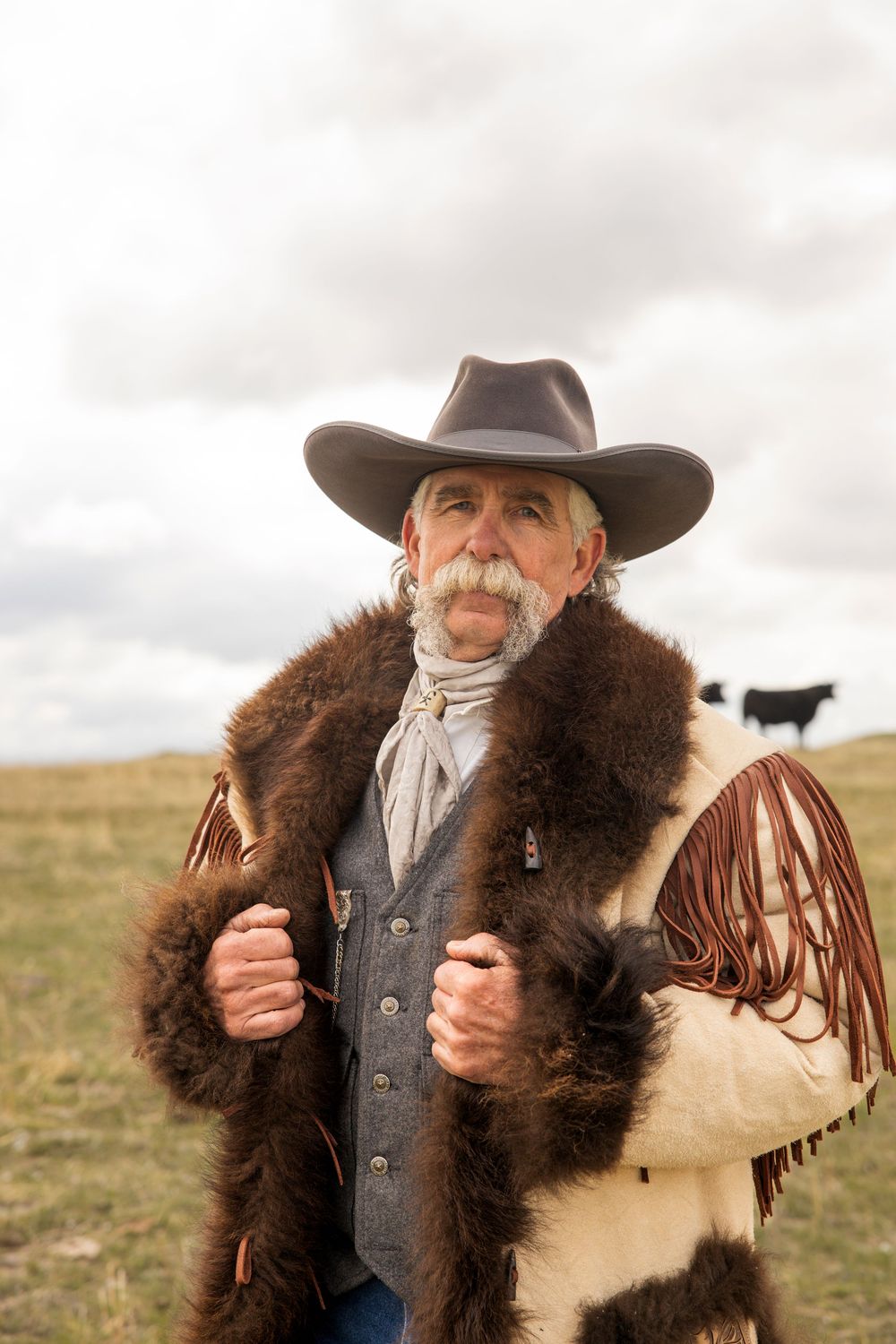 Cowboy wearing fur coat and western attire poses against cloudy winter landscape.
