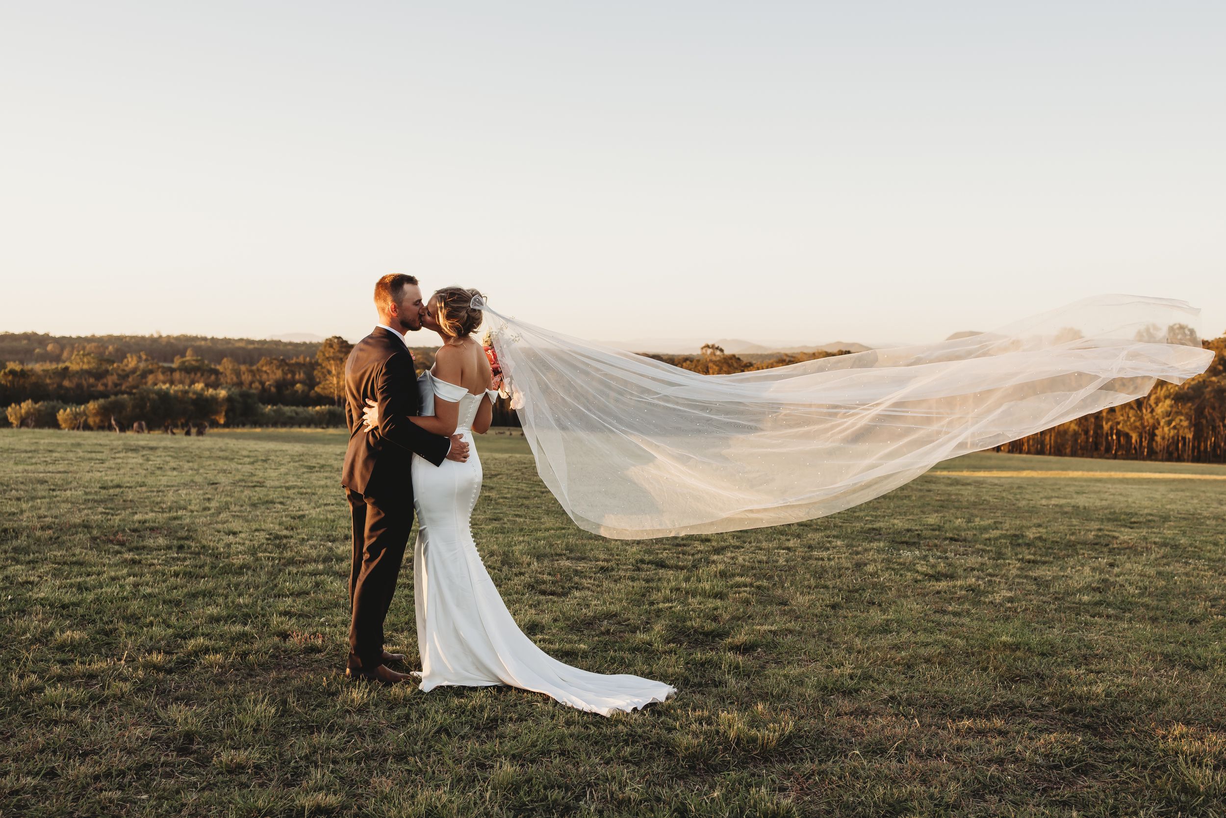 Bride and groom kissing in a field at sunset with wedding veil flowing dramatically in the wind.