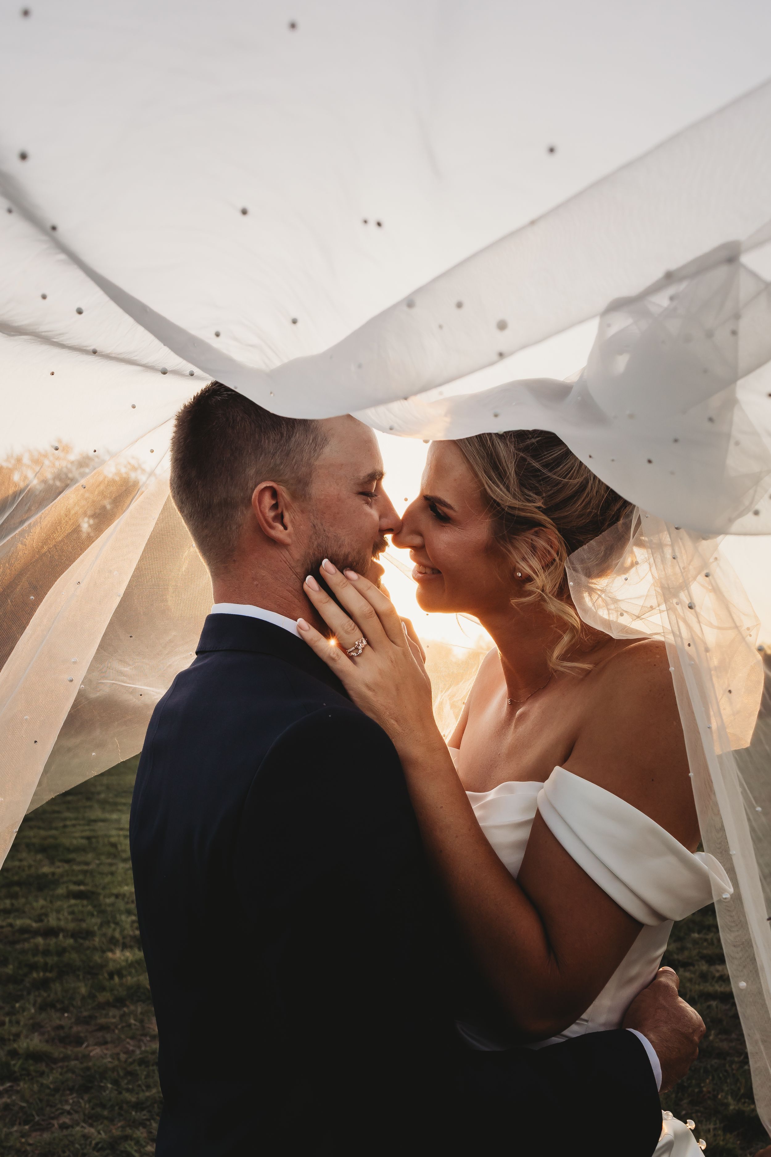 Bride and groom nearly kissing under flowing veil at sunset, bride tenderly holding grooms face.