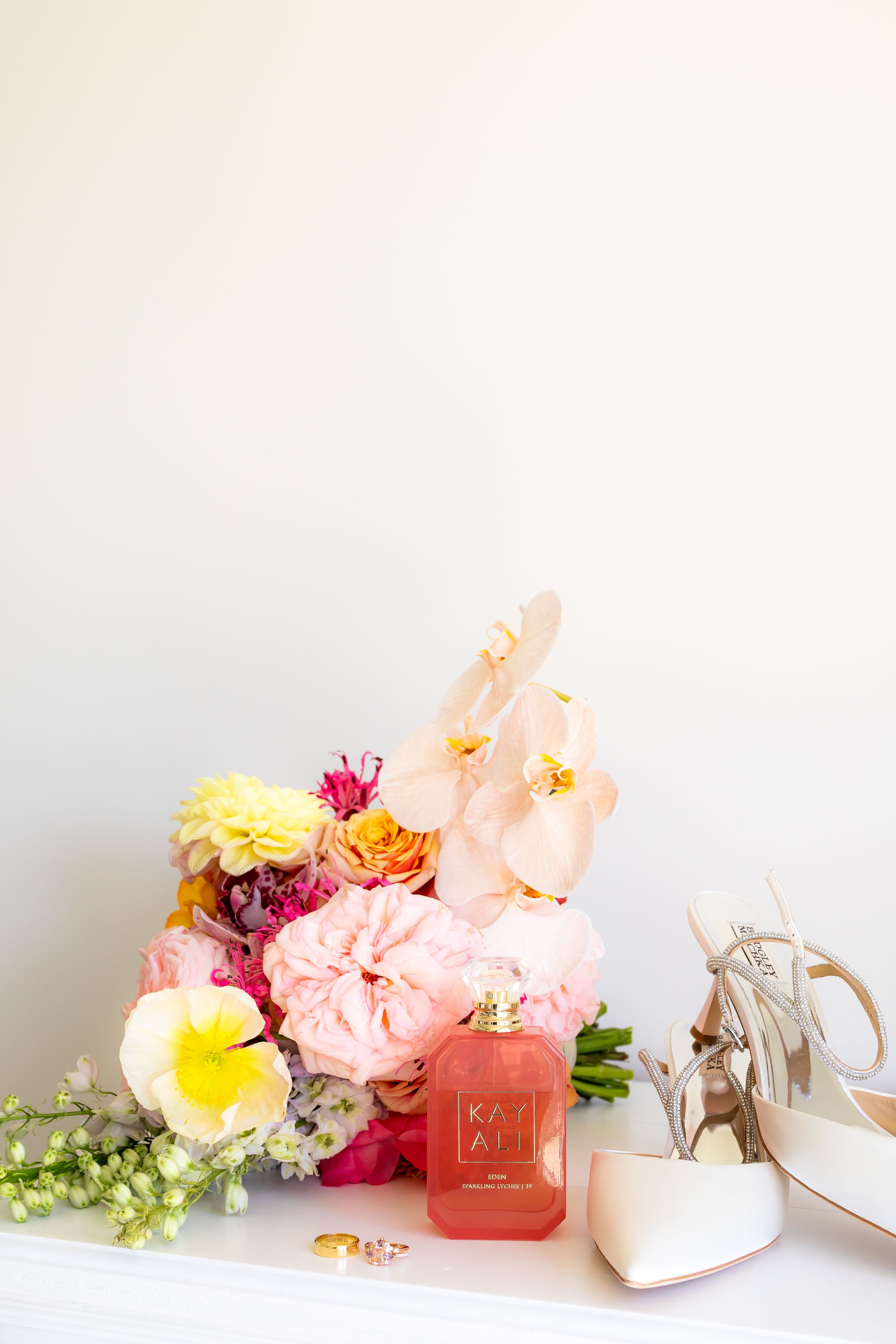 Bridal flat lay with colorful floral bouquet, coral perfume bottle, gold rings, and nude heeled sandals on white surface.