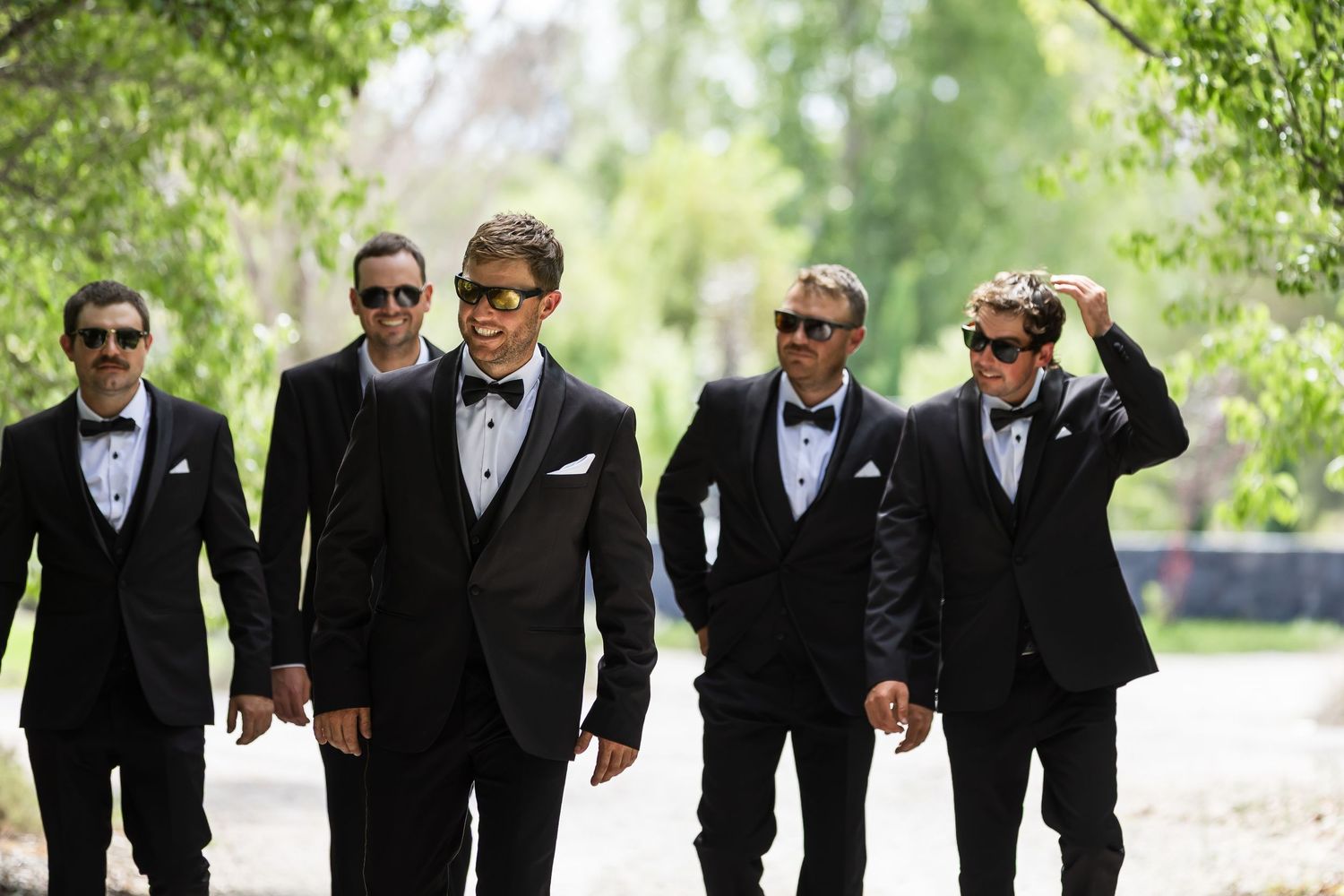 Six groomsmen in black tuxedos and sunglasses walking outdoors on a sunny day at a wedding.