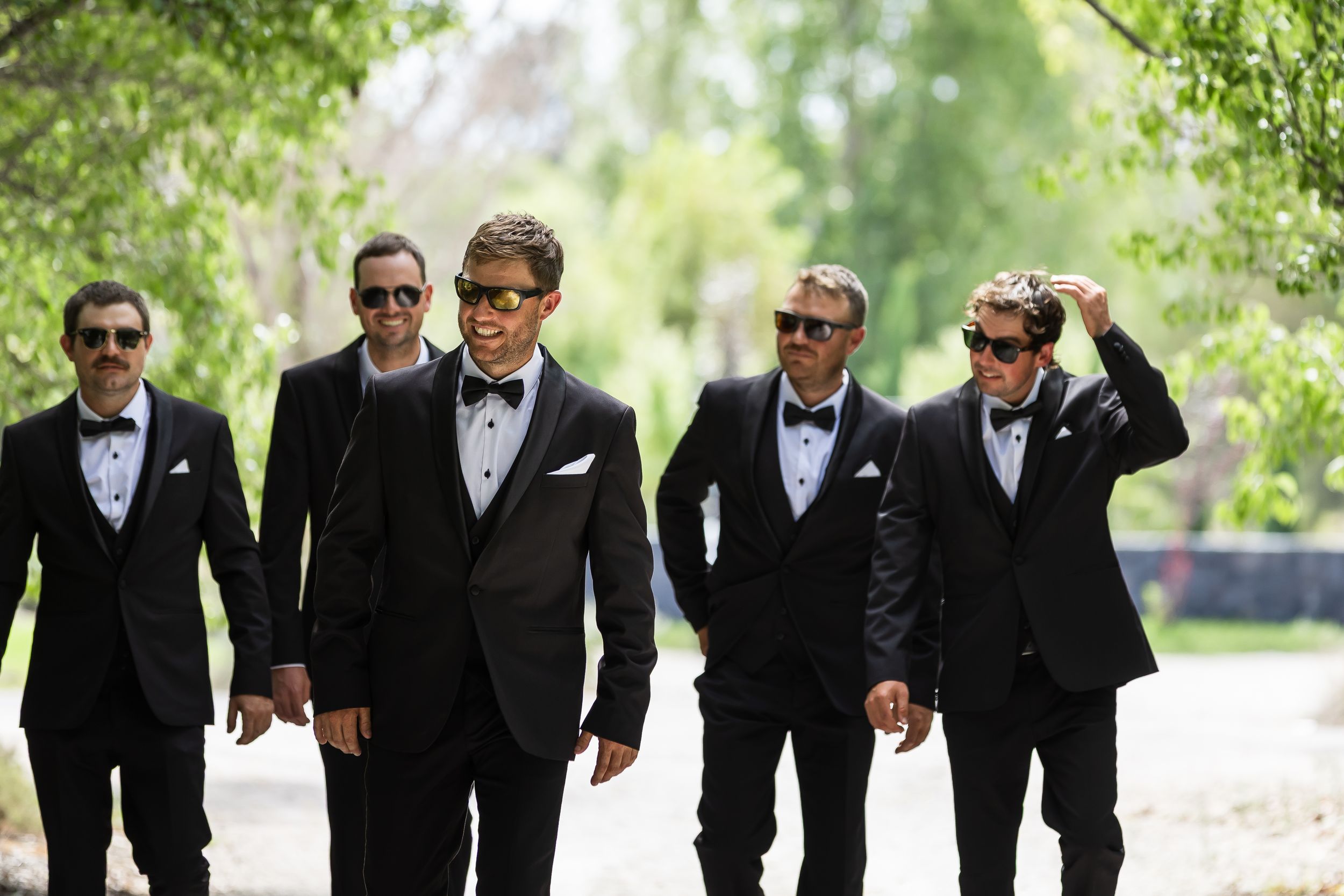 Six groomsmen in black tuxedos and sunglasses walking outdoors on a sunny day at a wedding.