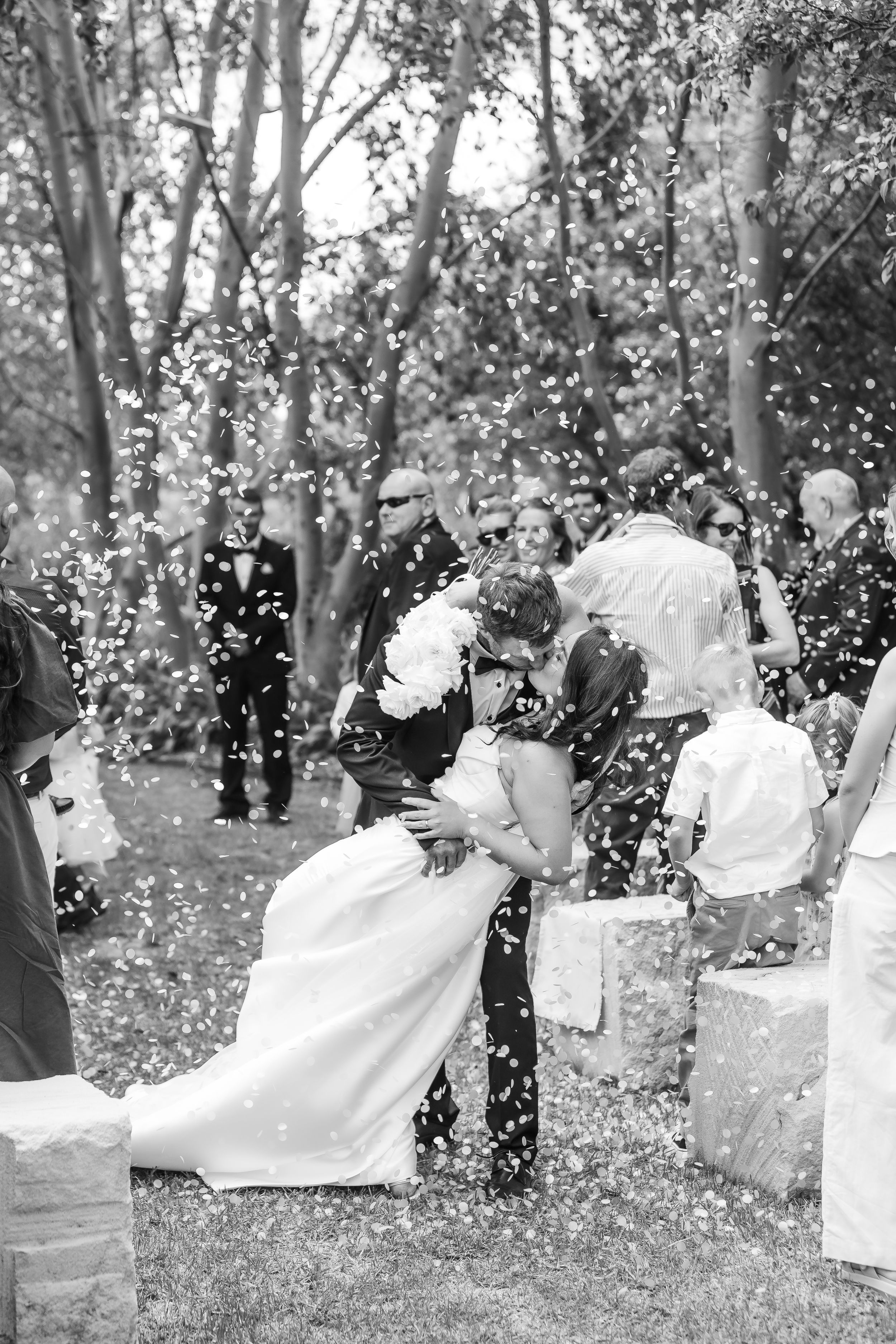 Black and white photo of bride and groom sharing a kiss as confetti falls during outdoor wedding ceremony.
