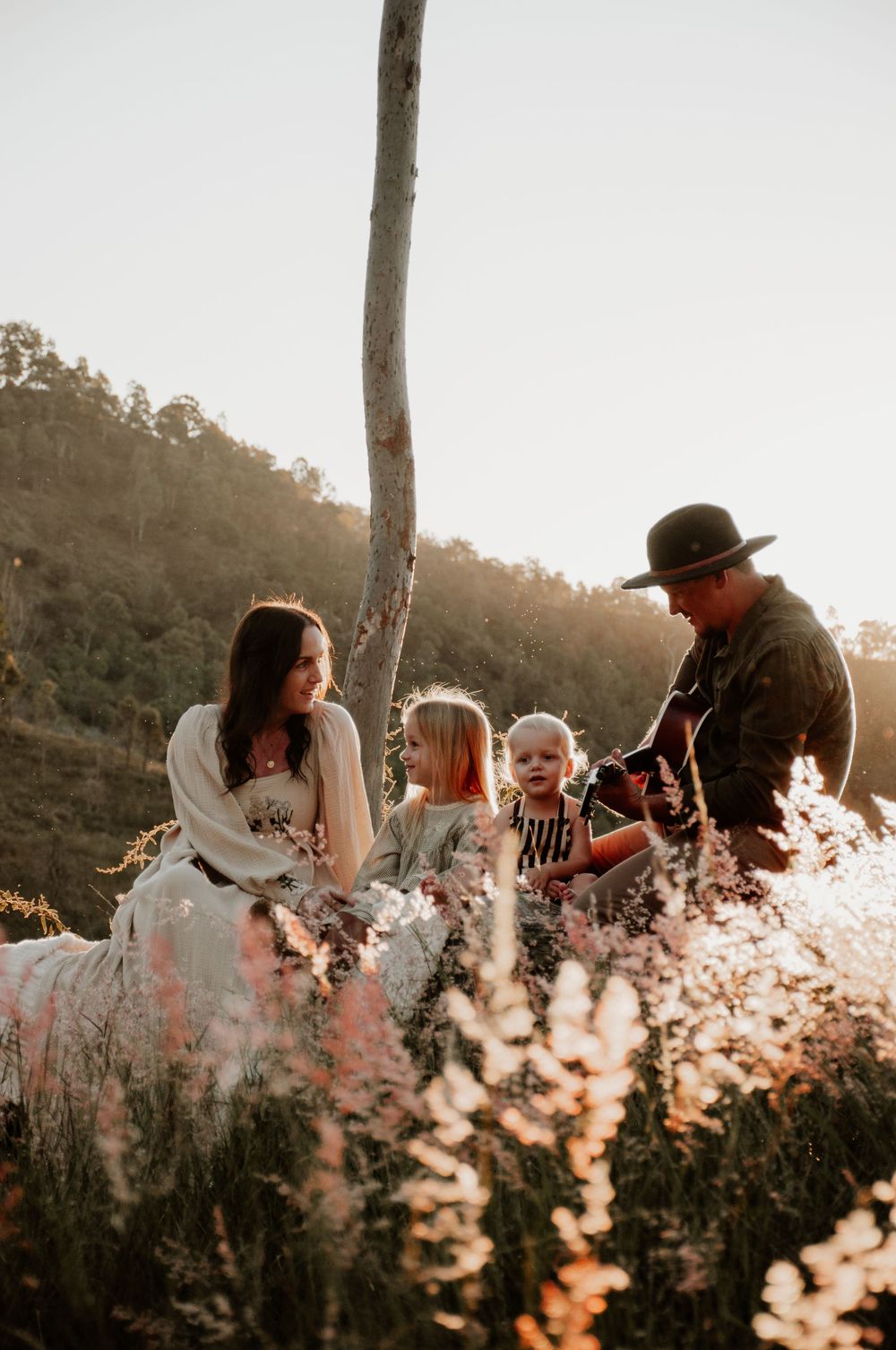 family sitting on a log in a field at sunset. dad plays the family guitar while they listen in