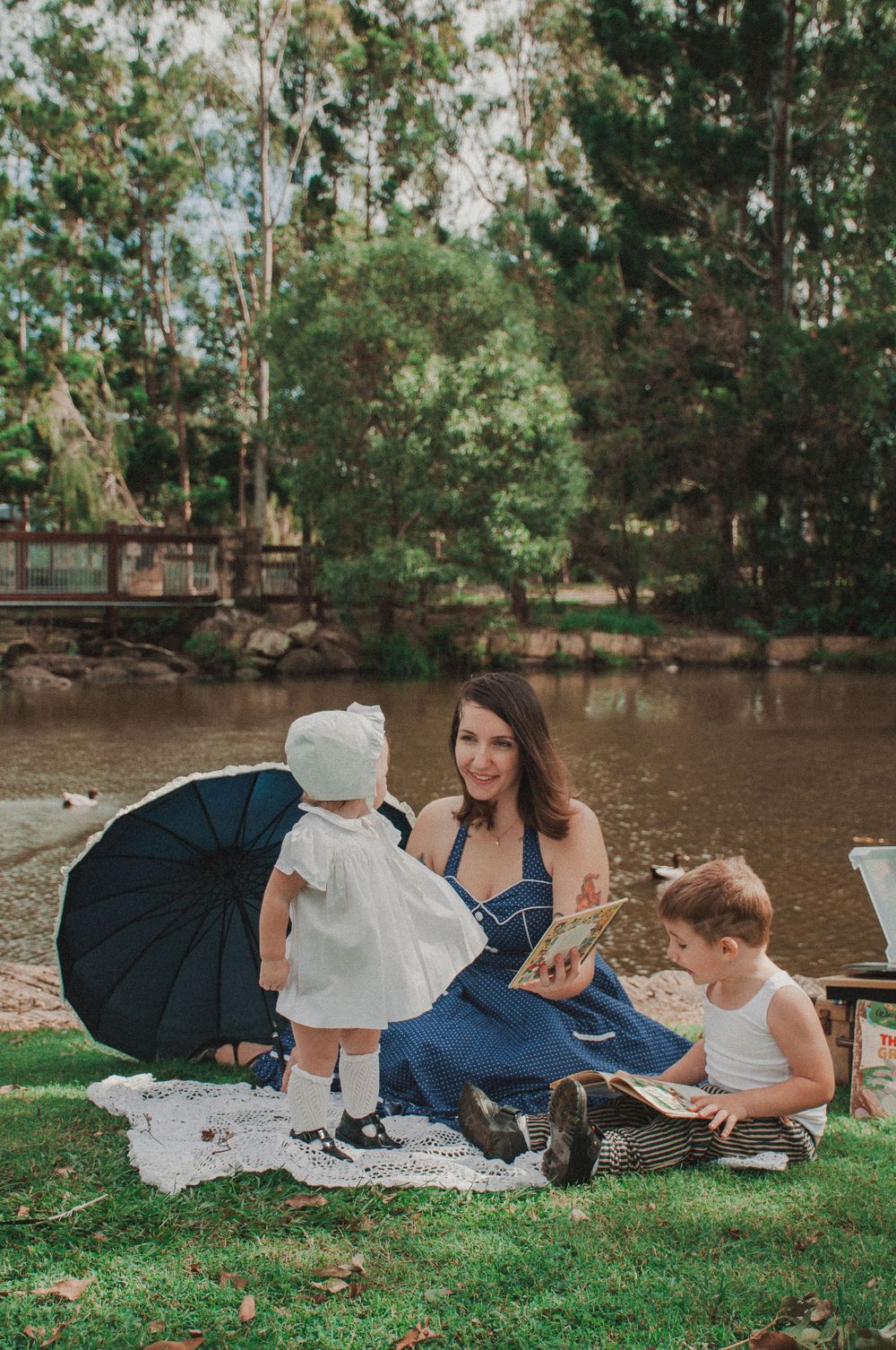 old style, retro style mum and bub photoshoot. mum and daughter and son read under an old umbrella in front of a duck pond