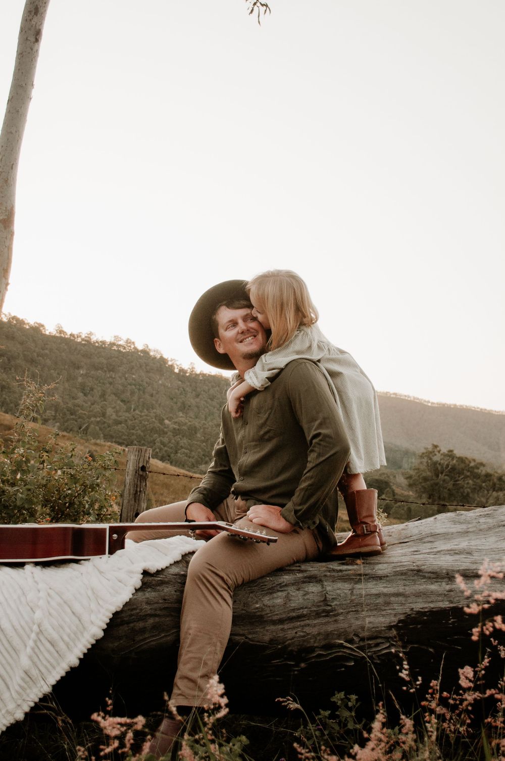 Daughter hugging dad while he plays guitar on a long in a field at sunset
