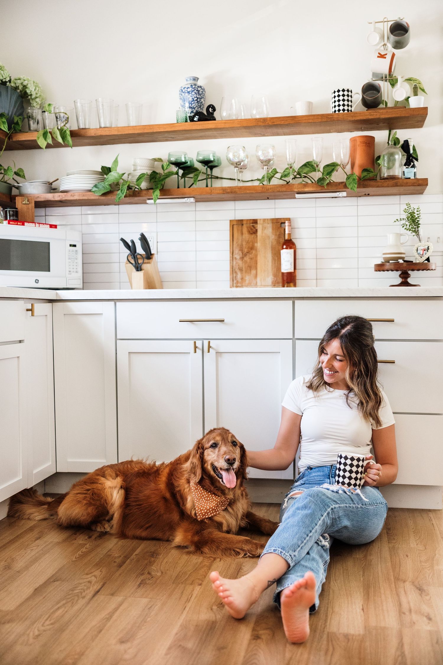 Young woman sits on kitchen floor with a golden retriever dog, holding a mug in a bright white kitchen.