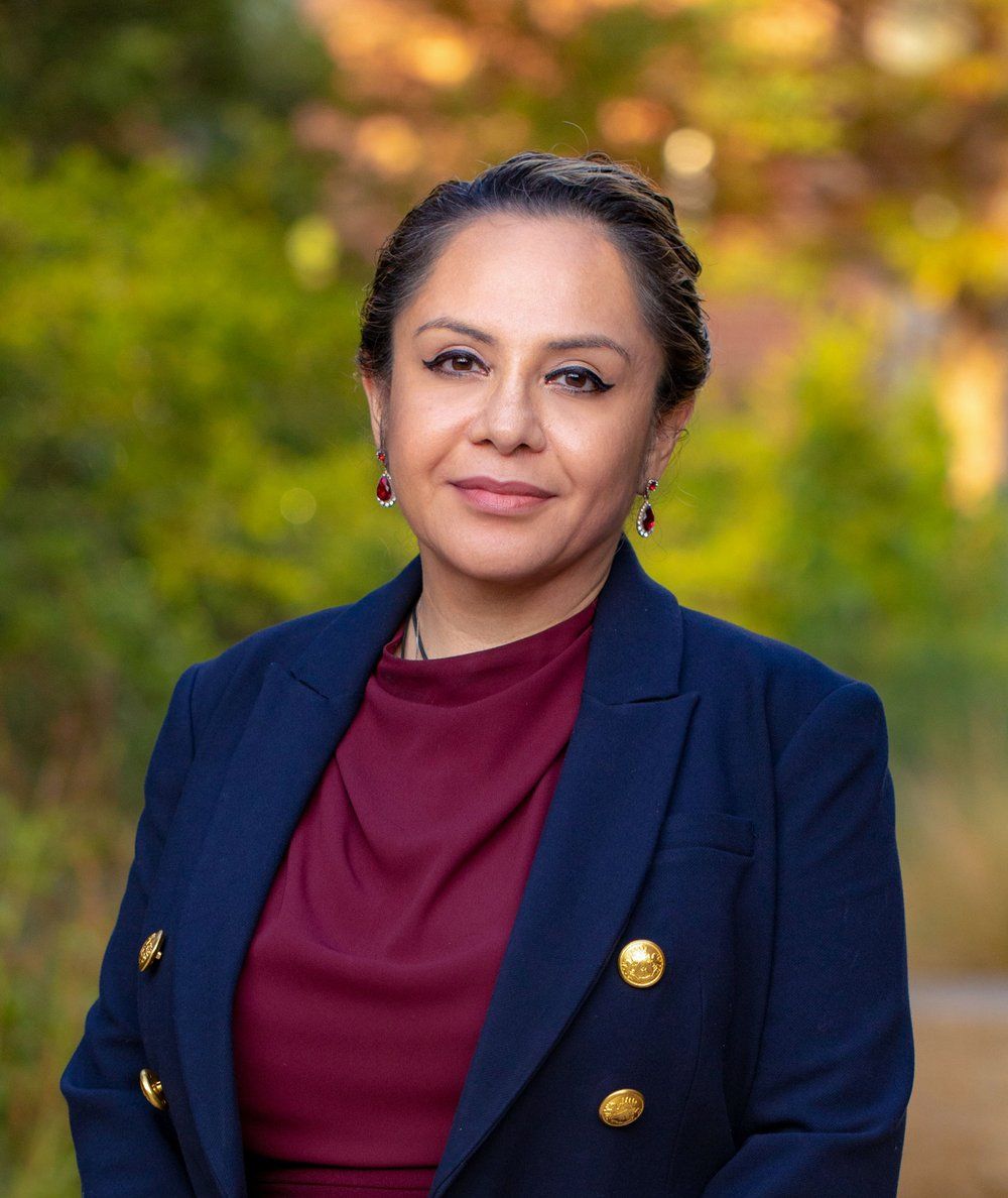 Professional headshot of someone in a navy blazer and burgundy top against an autumn background.