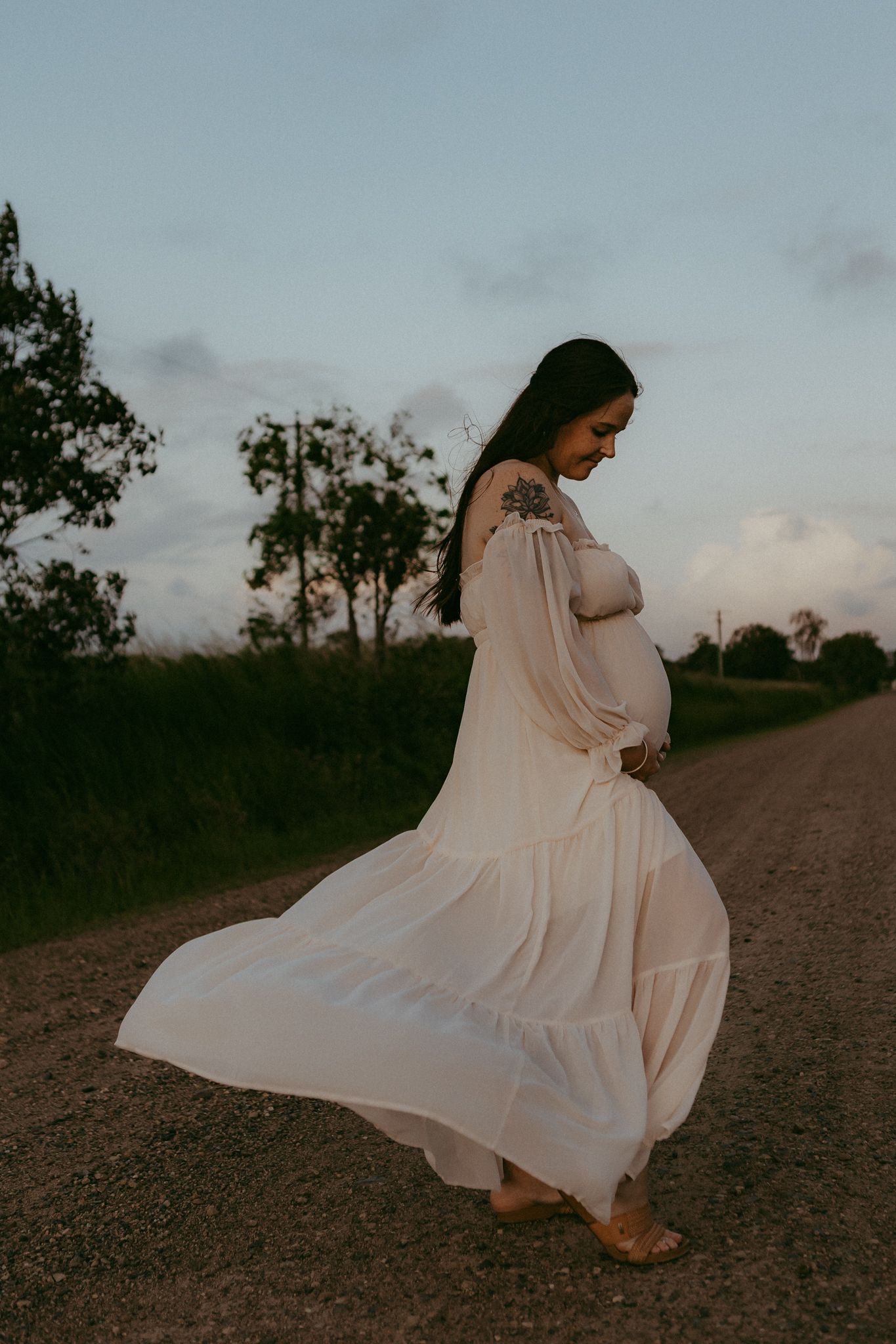 Pregnant woman in flowing white dress cradling her bump on a country road at twilight.