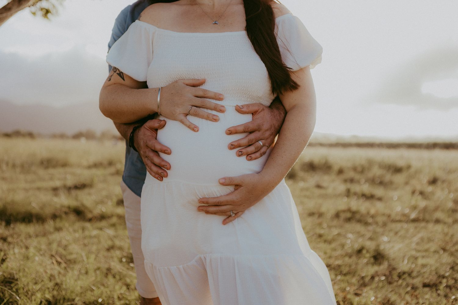 Pregnant woman in white dress with partner's hands cradling her baby bump outdoors at golden hour.