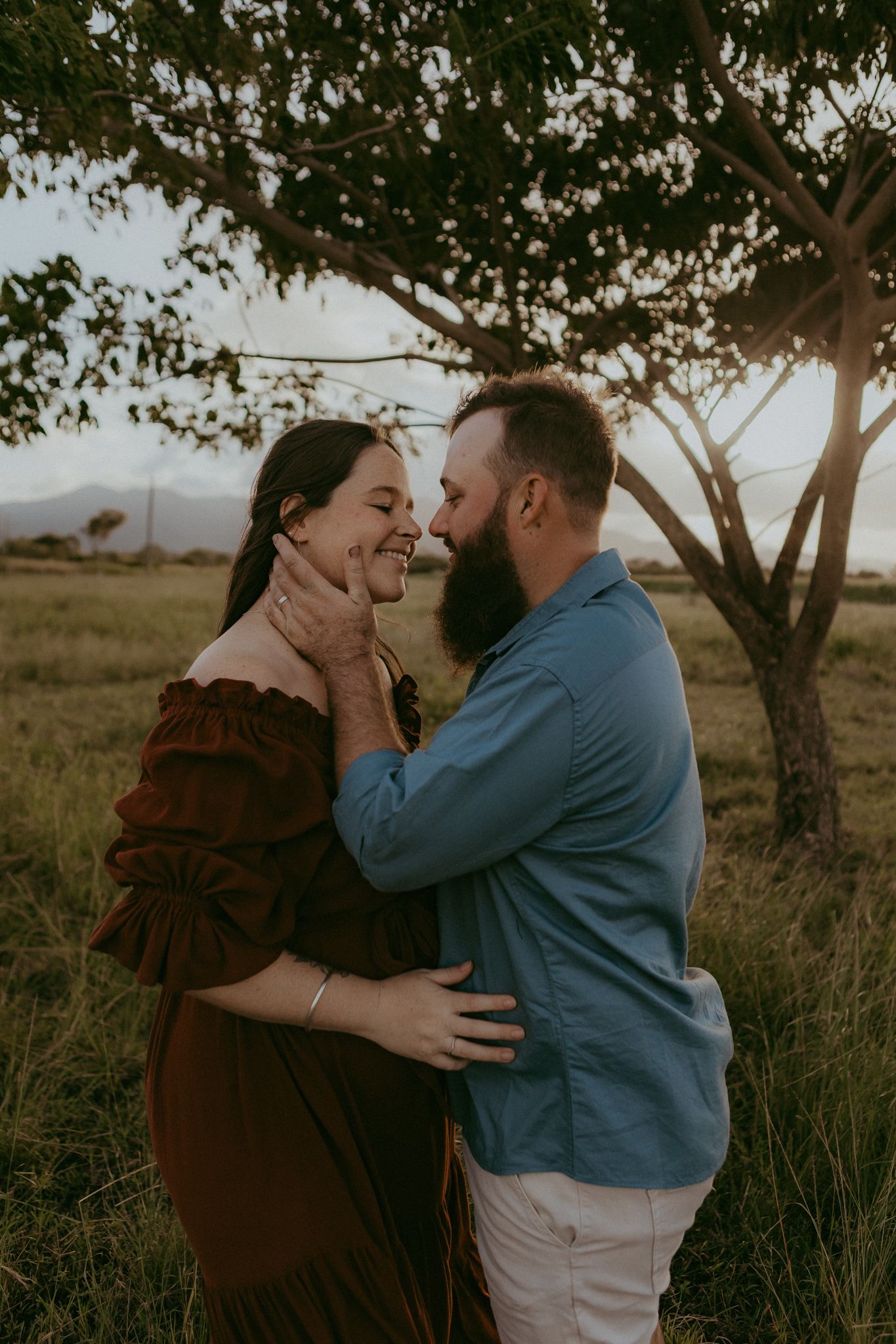 Bearded man tenderly holding pregnant woman's face as they smile at each other under a tree at sunset.