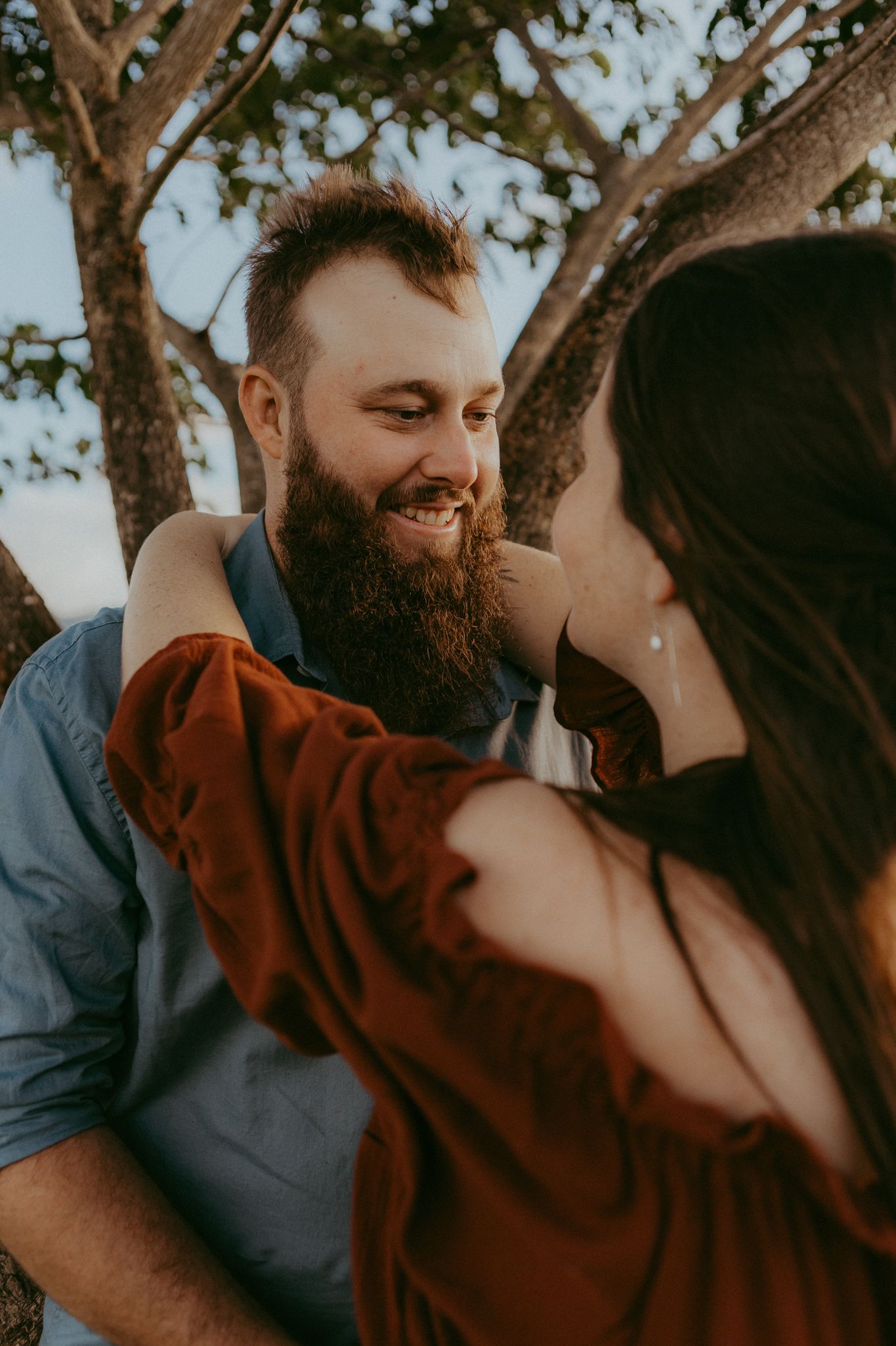 Smiling bearded man in blue shirt gazing at woman in rust dress during outdoor maternity photoshoot.